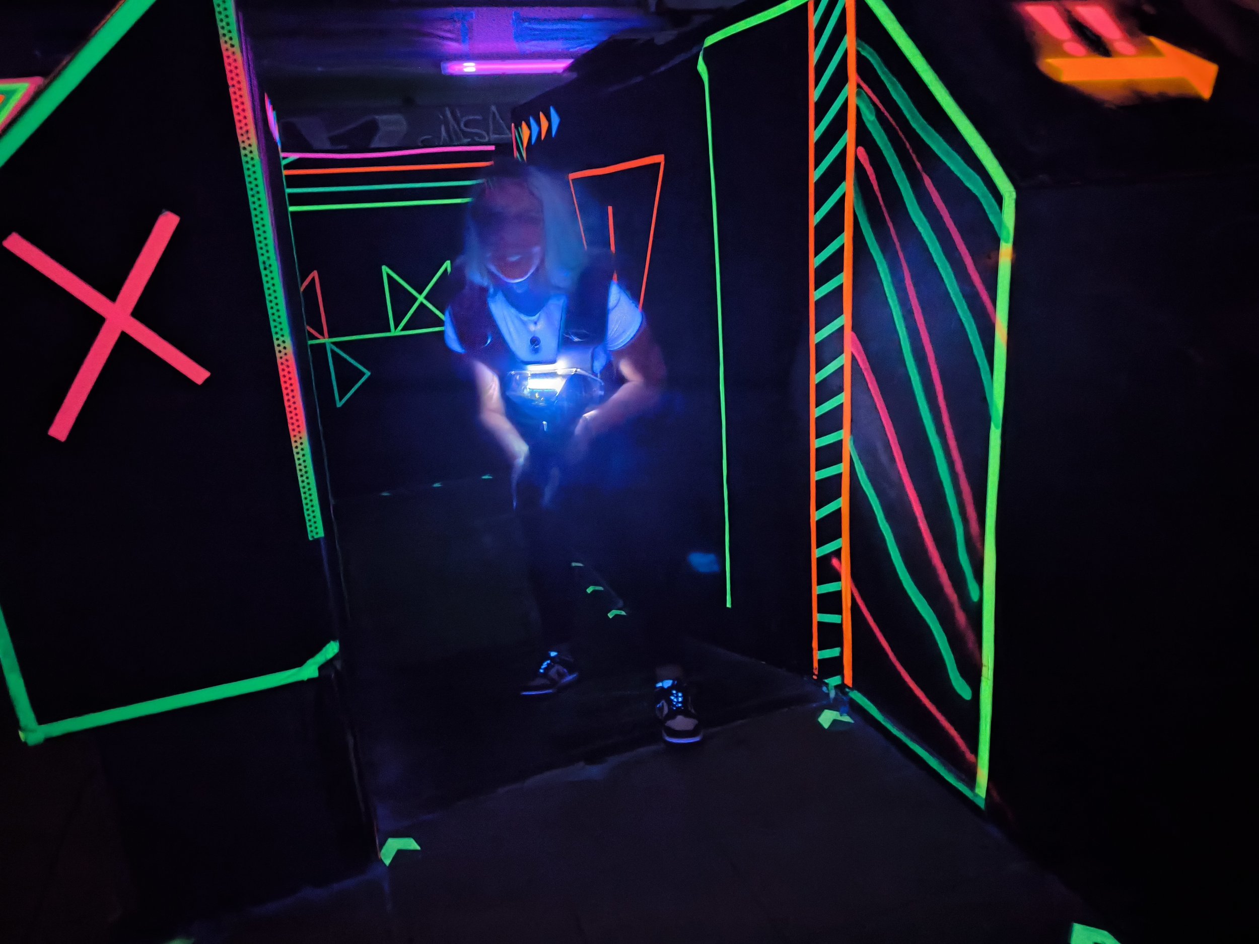 Eine Person in einer schwarzen Kleidung steht in einem Raum mit schwarz gestrichenen Wänden, die mit leuchtenden, neonfarbenen Linien und geometrischen Mustern bemalt sind, um eine futuristische Atmosphäre zu schaffen.