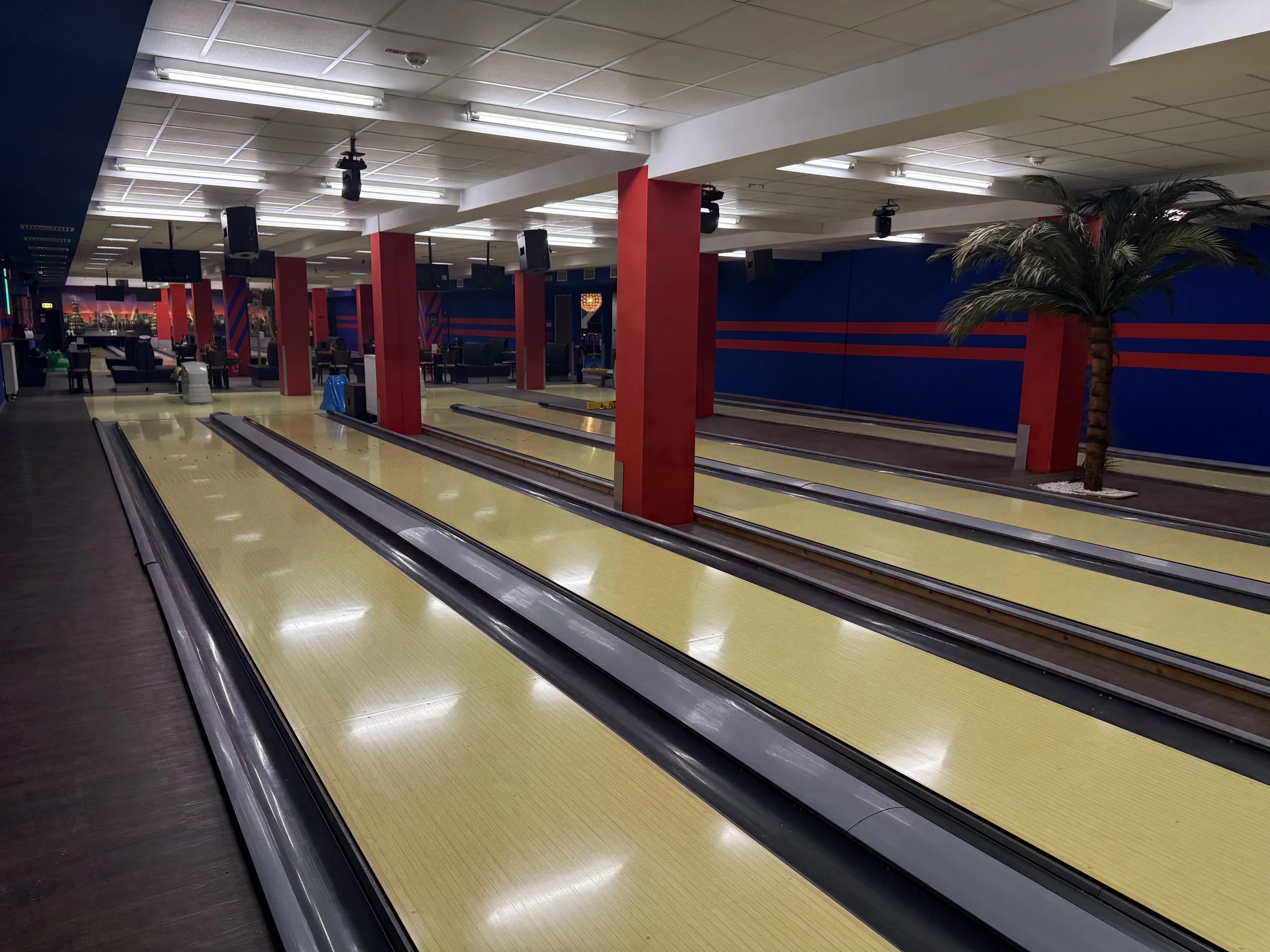 Leeres Bowlingcenter mit vier Bahnen, roten Säulen, einer Palme und Sitze im Hintergrund.