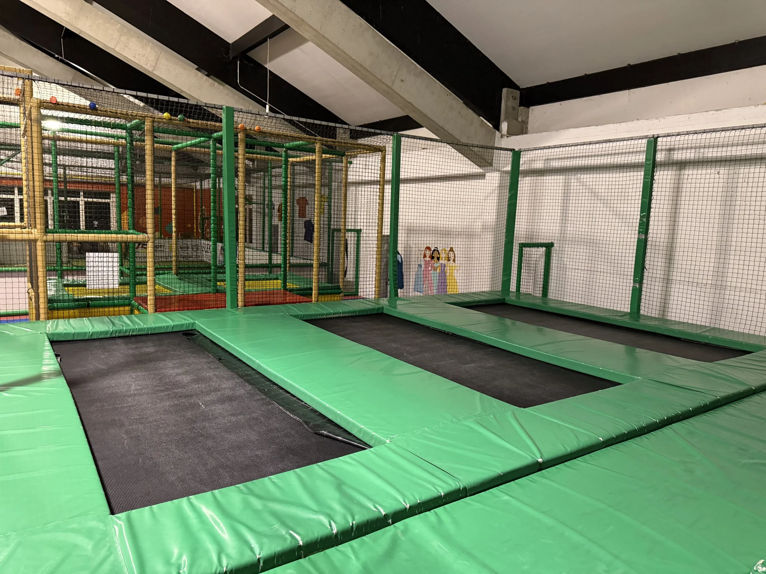 Innenaufnahme eines Spielplatzes mit mehreren Trampolinen und Klettergerüsten, umgeben von einem Netz.