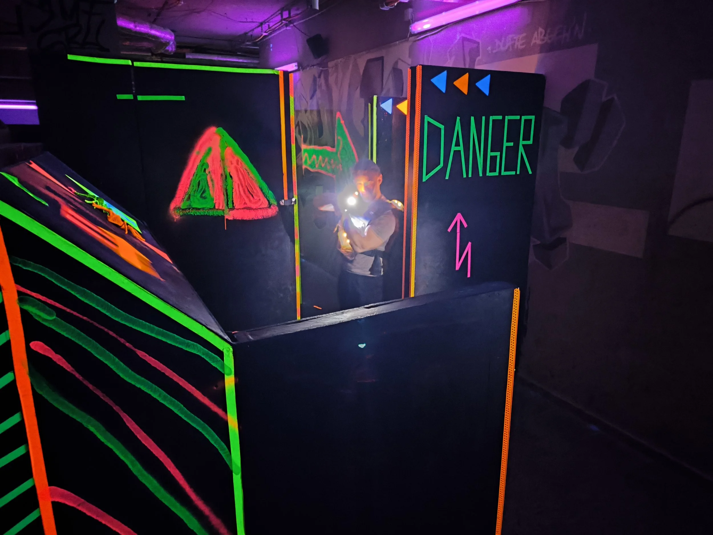 Eine Person steht in einer dunklen, mit Neonfarben beleuchteten Raum, hinter einer Barriere mit einer Warnung "DANGER". Die Neonzeichen zeigen ein Blutdruckdiagramm, ein Warnschild und Symbole in Neonfarben.