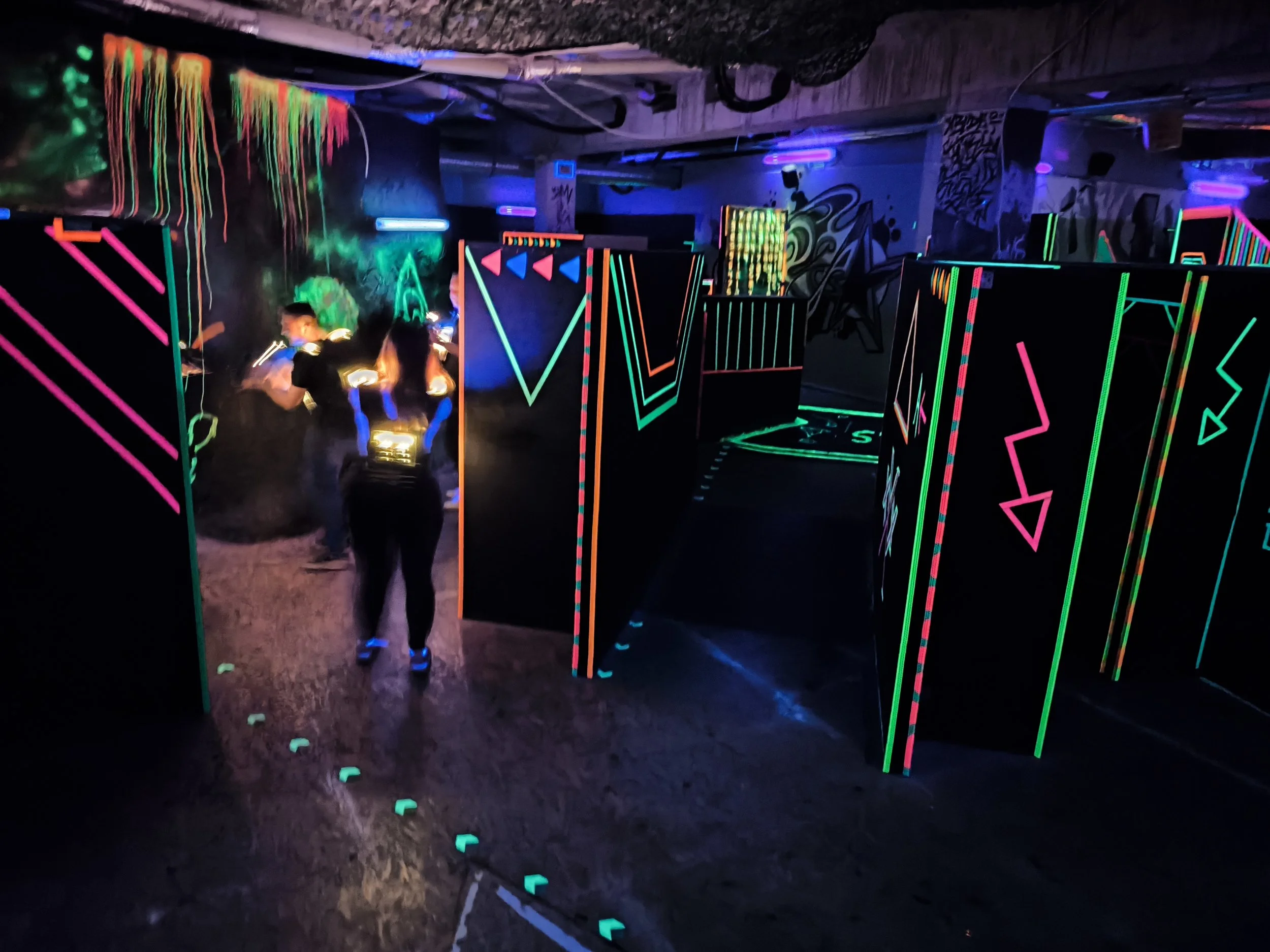 Dunkel beleuchtener Raum mit Neonfarbenen Linien und Figuren, Menschen beim Spielen eines Trampolins, Neon-Kunst und Graffiti an der Wand.