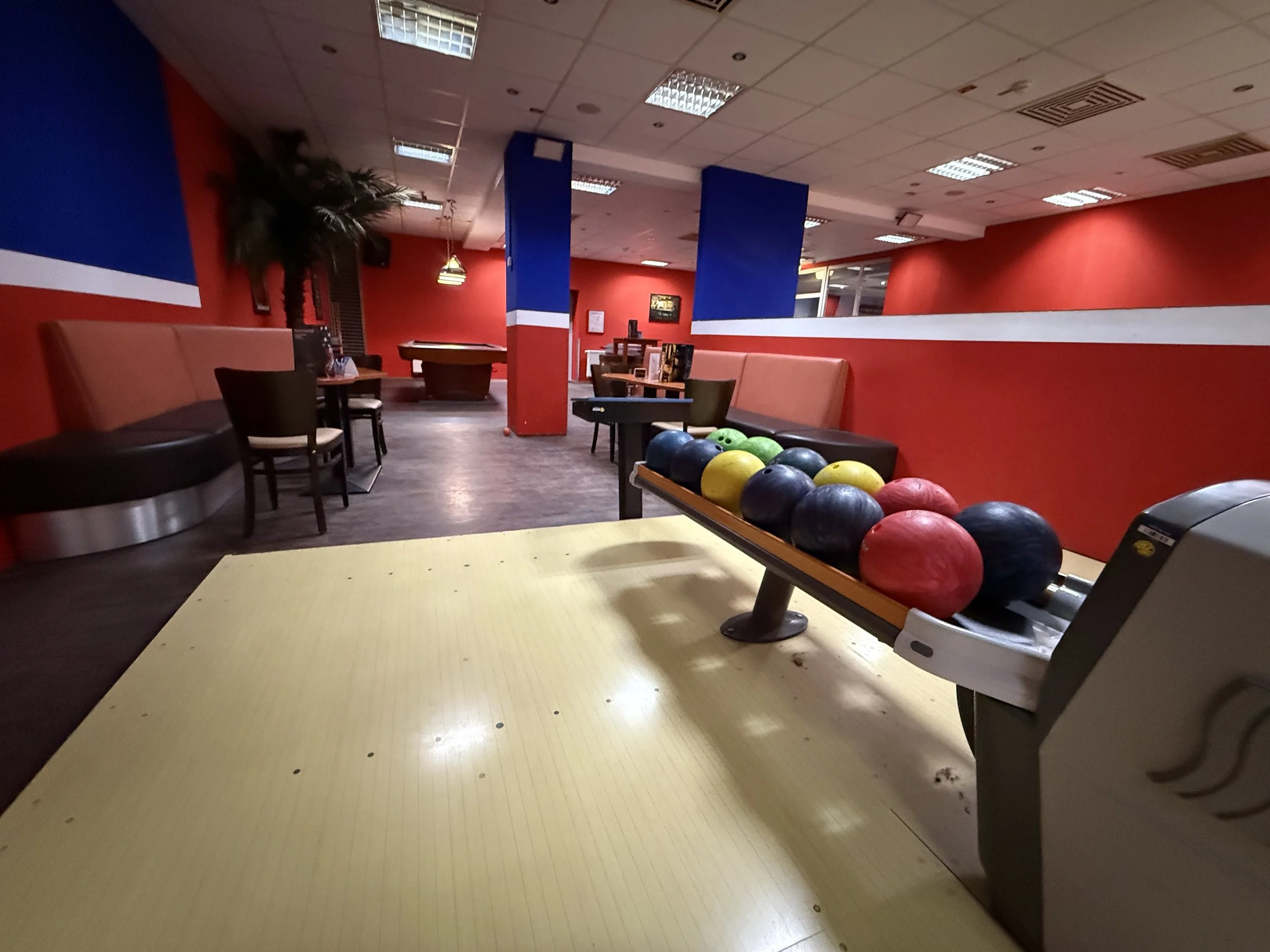 Leerer Bowlingbahnhof mit mehreren bunten Kugeln auf der Kugelrolle, rote, blaue und braune Sitzgelegenheiten, rote Wände, Deckenleuchten, grauer Boden, im Hintergrund ein Billardtisch.