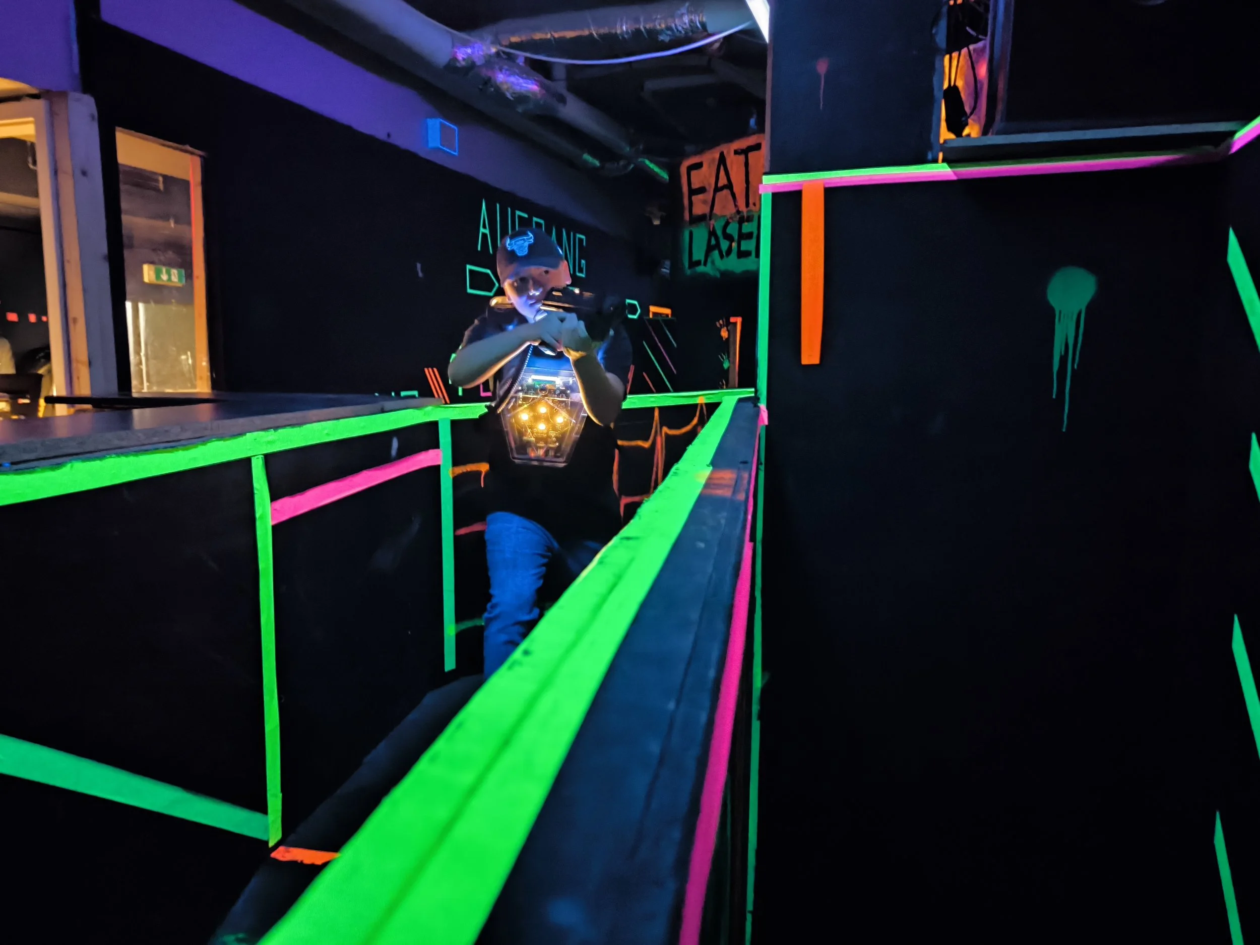 Person mit Neonfarben bekleidet in einer Schwarzlicht-Indoor-Arena bei Laser-Tag, zielt mit einer Waffe, im Hintergrund leuchten Neon-Schilder und -Grafiken.