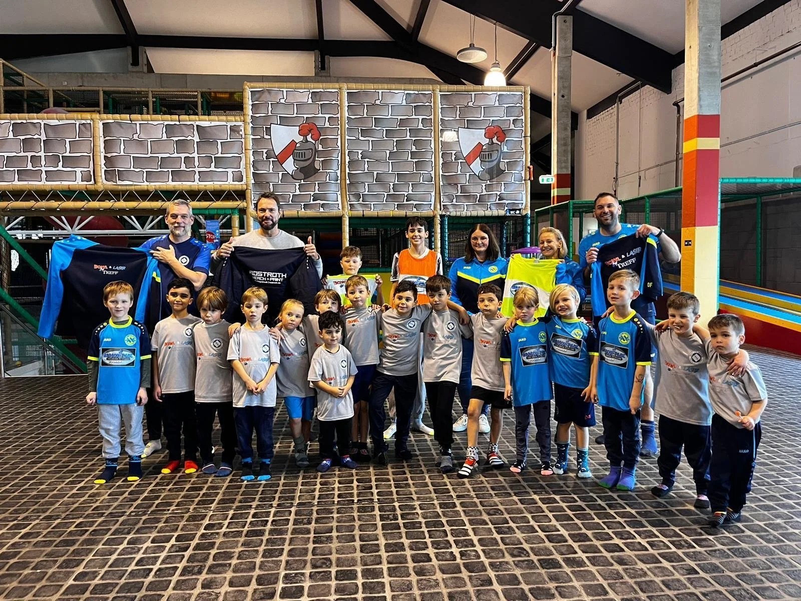 Gruppe von Kindern und Trainern in einer Indoor-Kletterhalle, meisten Kinder in grauen und blauen Trikots, einige halten Trikots hoch, im Hintergrund eine Kletterwand mit Burgmauer-Design.