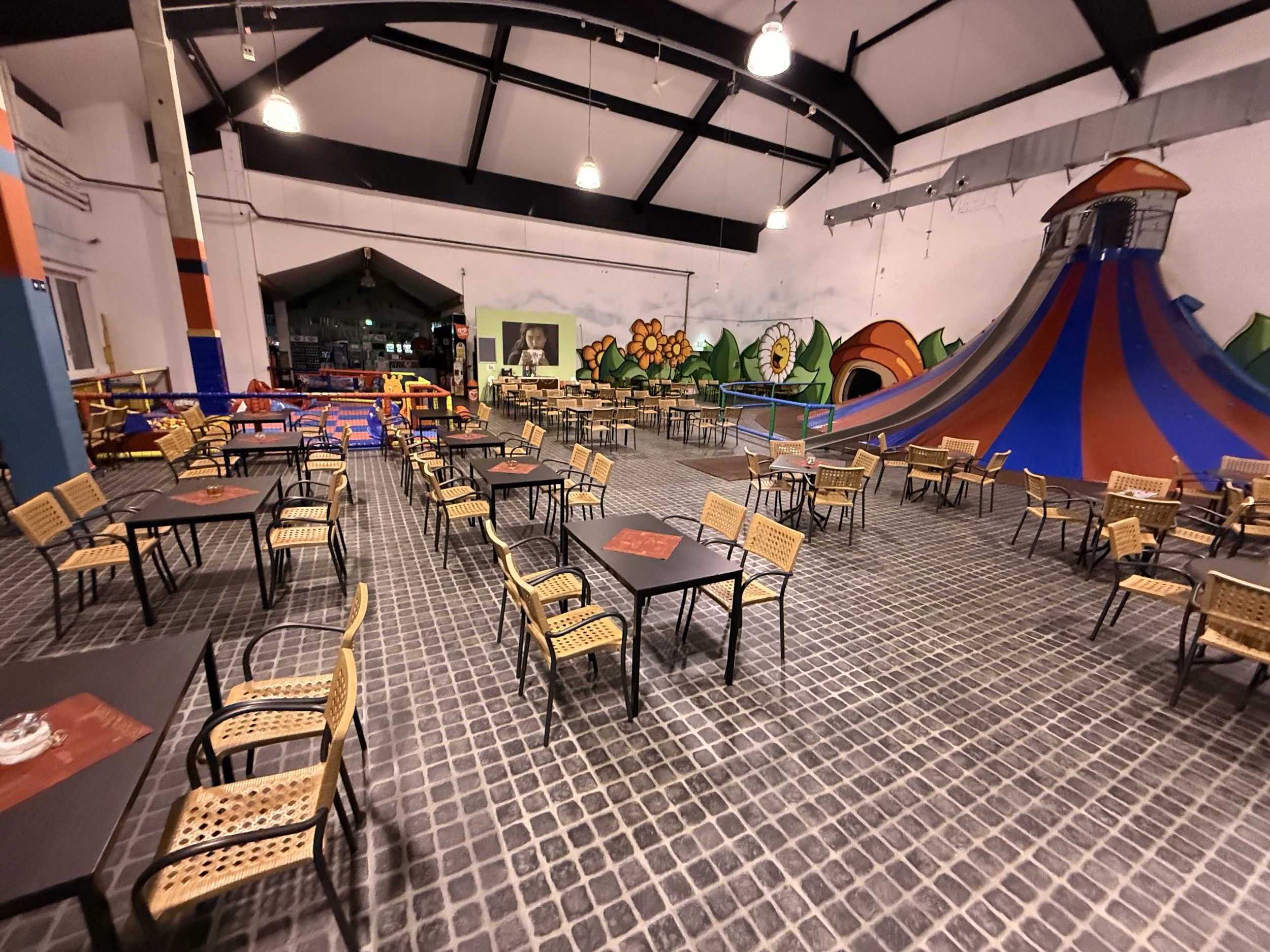 Blick in eine leere Indoor-Kinder-Spielhalle mit Sitzbereichen, buntem Rutschenturm und Wanddekoration mit Blumen. Es gibt Tische und Stühle, Luftballons und eine Attraktion, die an ein Zirkuszelt erinnert.