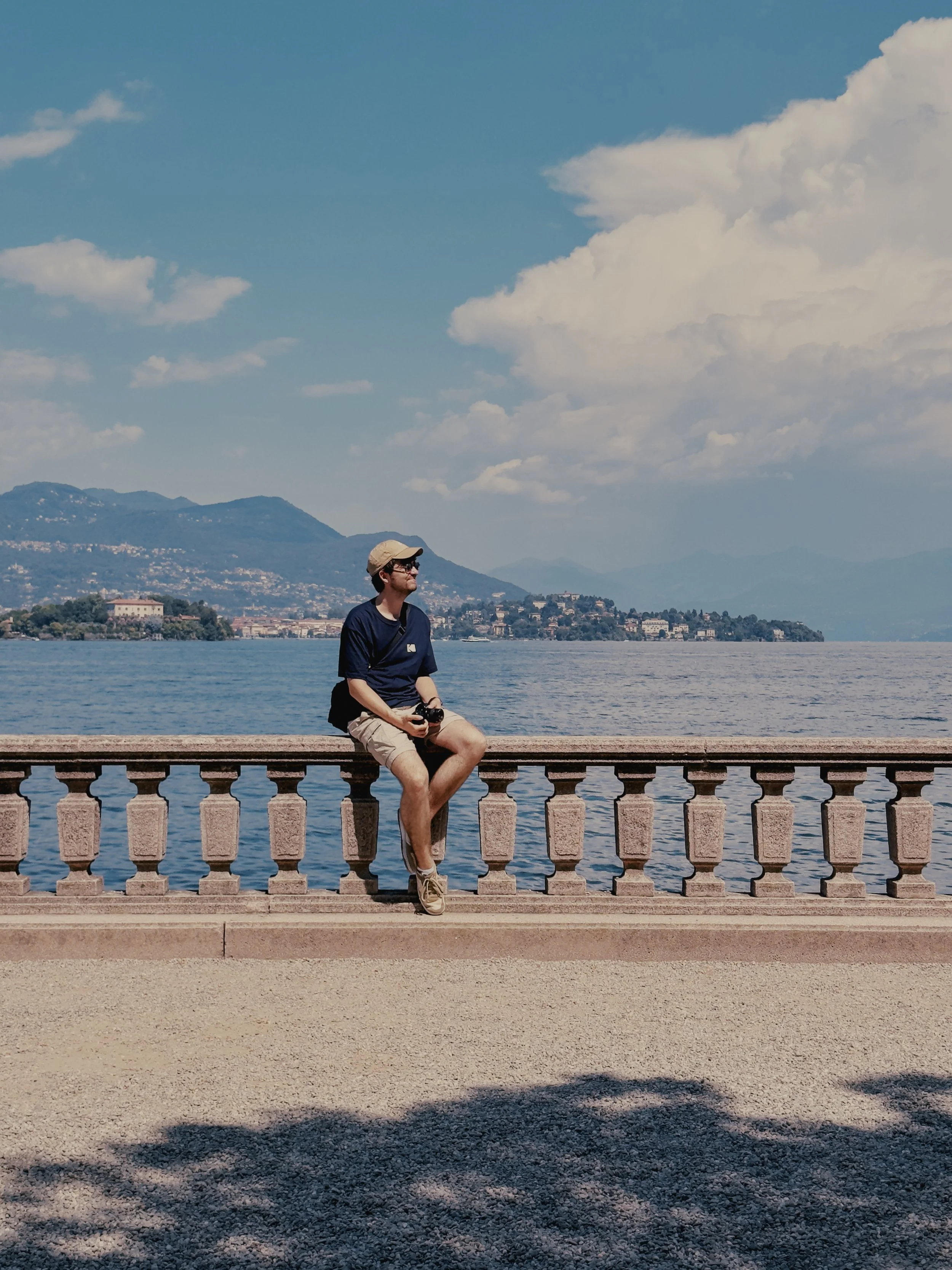 Man zit op een sierlijke stenen reling langs een meer of groot water, met bergen en een stad in de achtergrond, onder een deels bewolkte blauwe hemel.