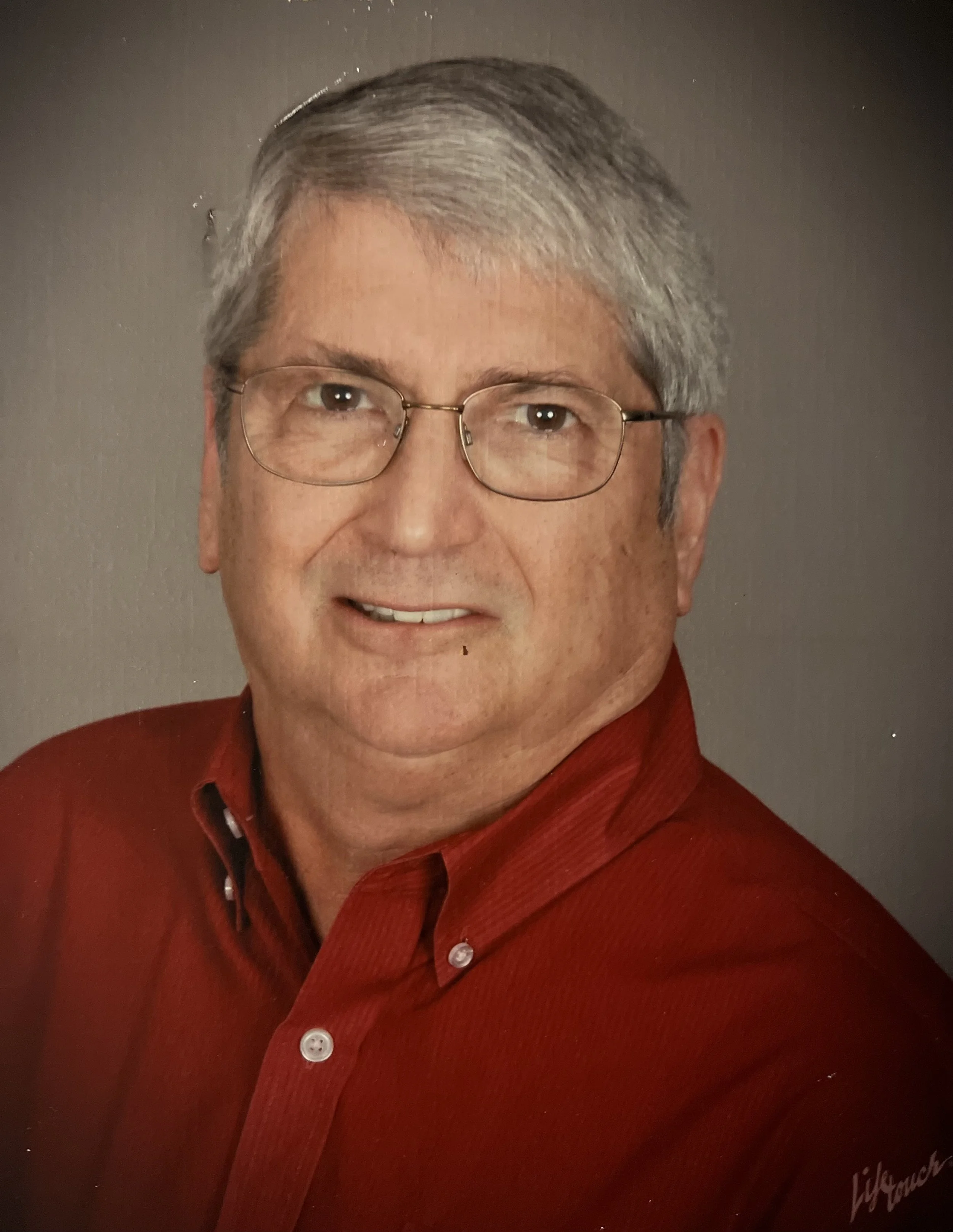 Portrait of an older man with gray hair, wearing glasses and a red shirt, smiling at the camera.