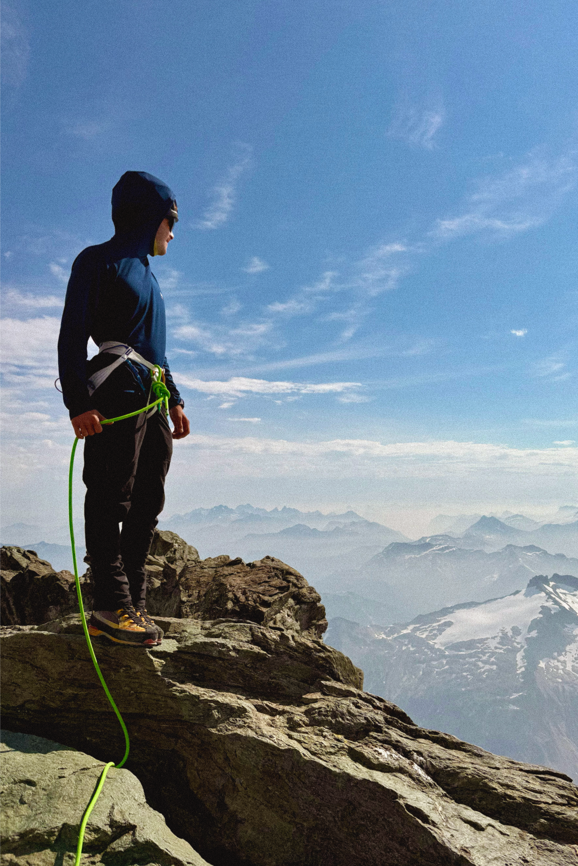 Ben standing on a rocky mountain ledge, overlooking a mountain range with snow-capped peaks under a blue sky.