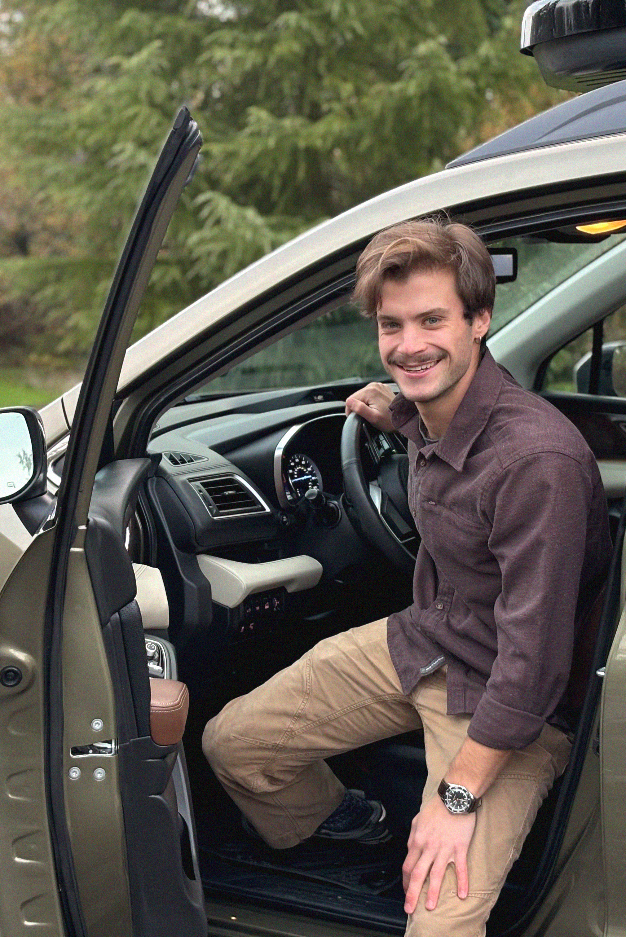 A man with brown hair, wearing a brown shirt and khaki pants, sitting in the driver's seat of a vehicle with the door open, smiling at the camera, with a background of green trees.
