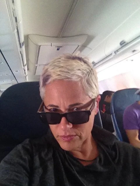 Person with blonde hair wearing black sunglasses sitting on an airplane.