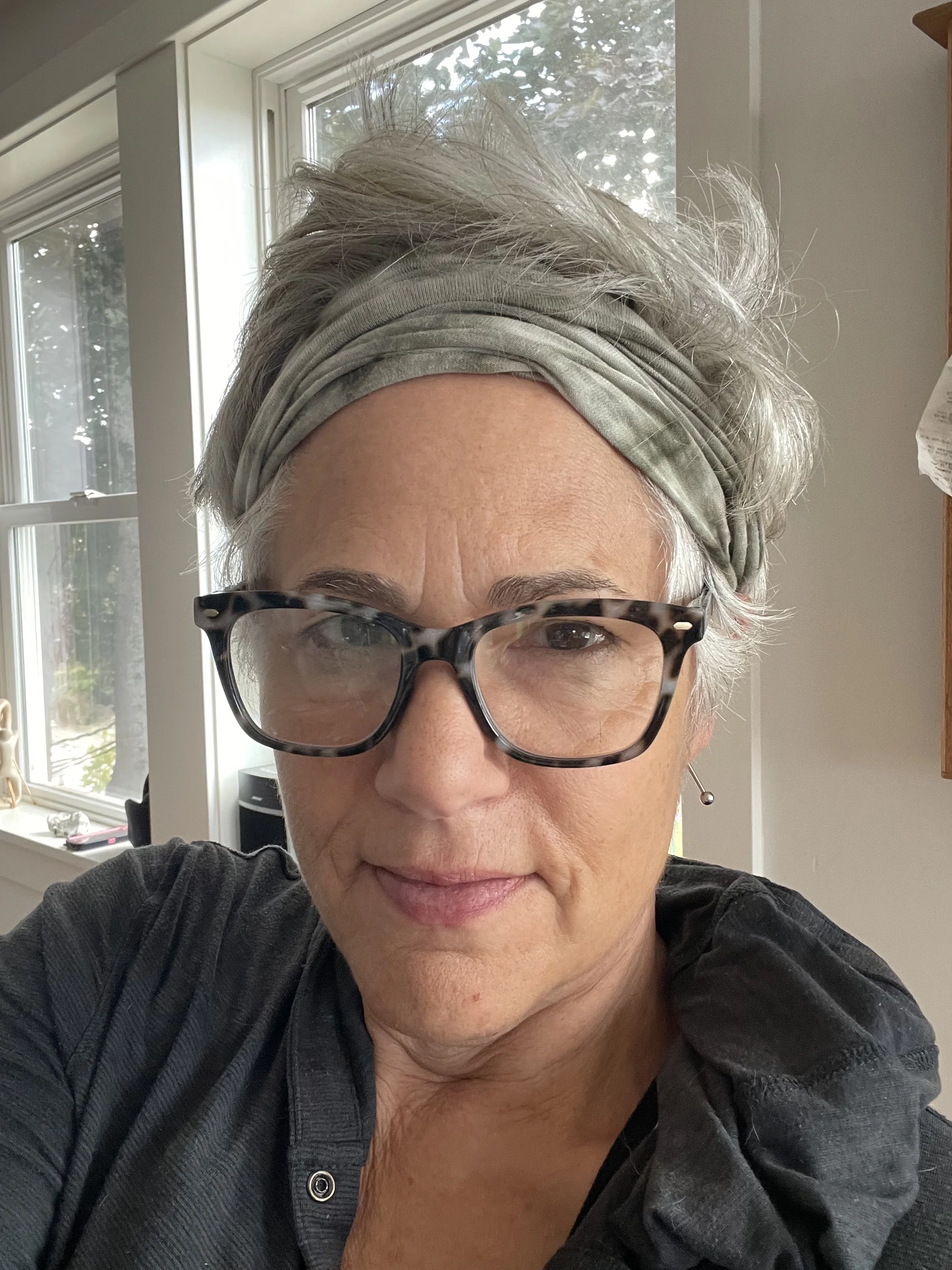 A woman with short gray hair, wearing a gray headband, large tortoiseshell glasses, and a dark shirt, is taking a selfie indoors near a window with natural light.