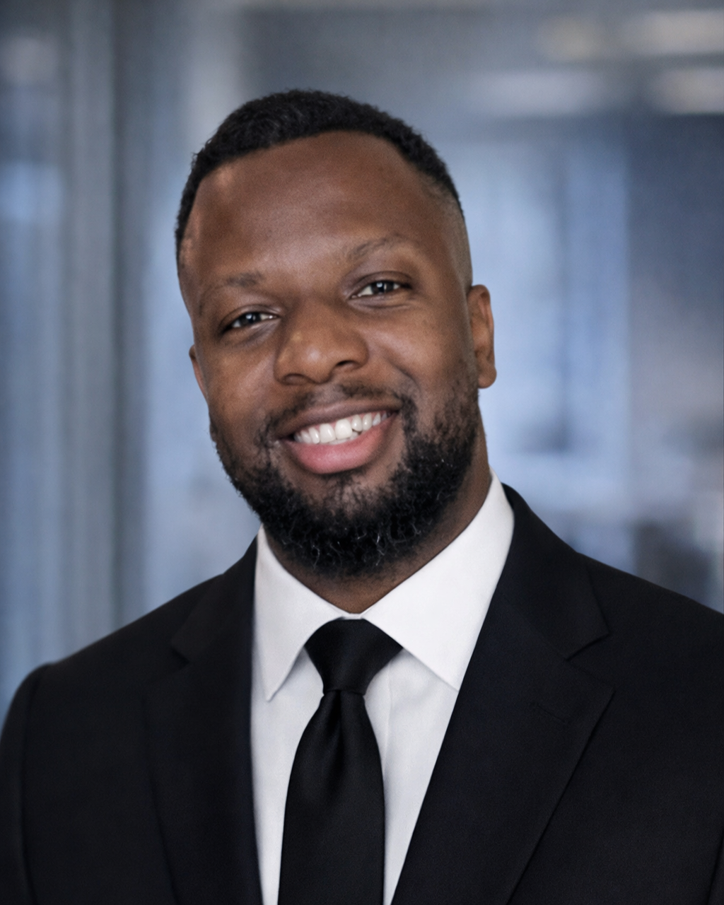 A smiling man in a black suit and tie, with short hair and a beard, posing against a blurred gray background.