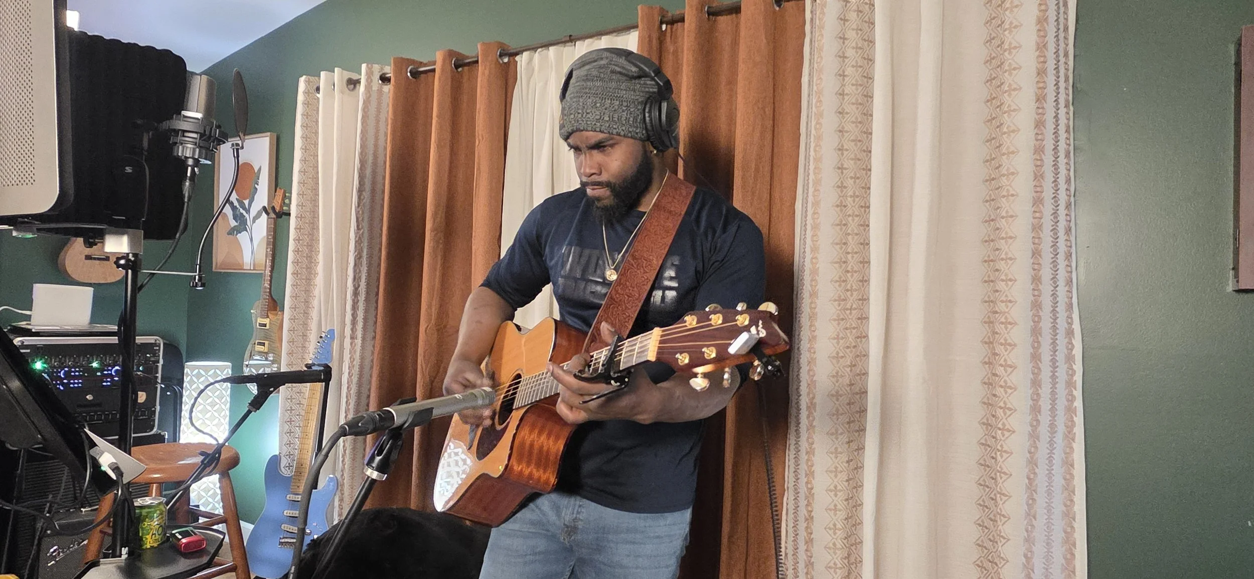 A man wearing a gray beanie, black t-shirt, and jeans playing an acoustic guitar in a room with musical equipment and guitars hanging on the wall.
