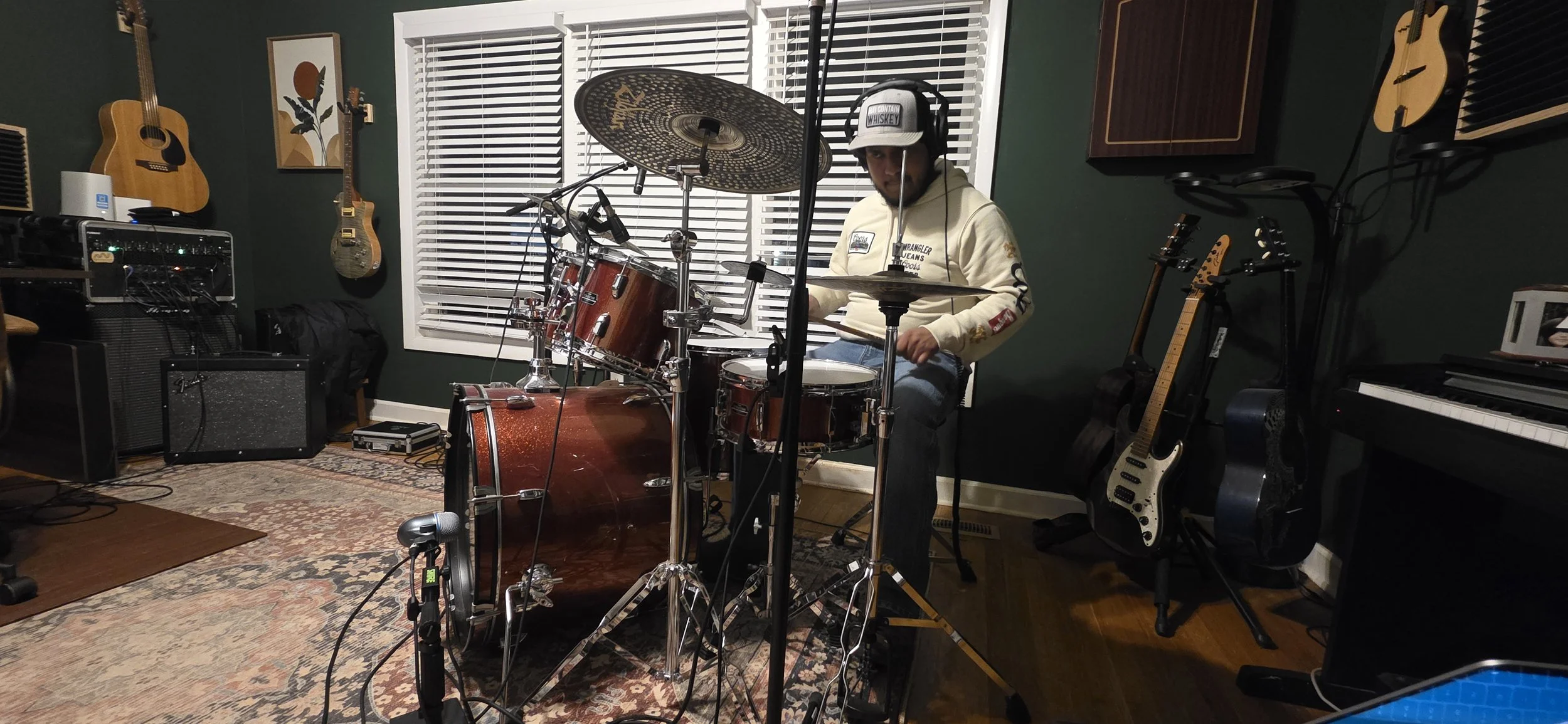 Man playing drum set in music studio with guitars hanging on wall and keyboard nearby.