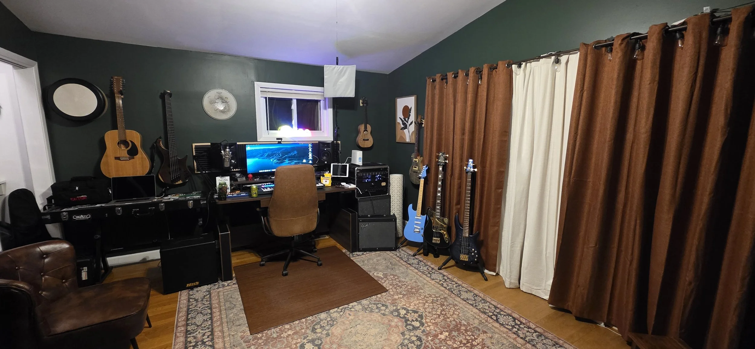 A music studio with dark green walls, equipped with guitars hanging on the wall, a computer, microphones, and other music gear, and brown curtains covering a window.