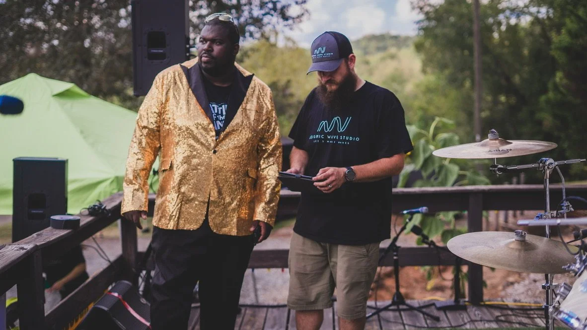 Two men standing outdoors on a wooden deck, with music equipment including drums and speakers around them. One man wears a shiny gold blazer and black pants, the other in a black t-shirt and khaki shorts, both focused on something.
