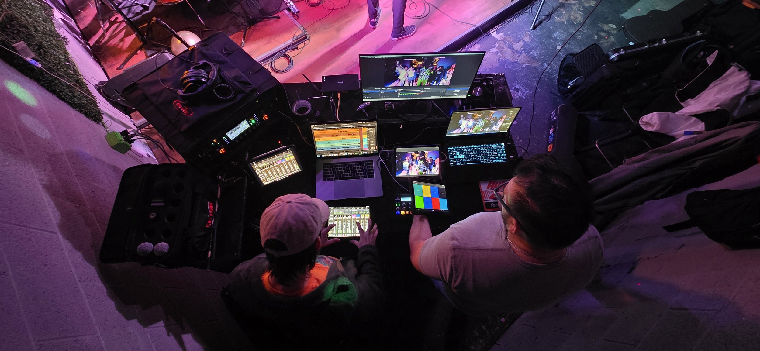 Overhead view of two people, a child and an adult, working at a DJ or live event setup with multiple laptops, tablets, monitors displaying colorful visuals, and audio equipment, with colorful stage lighting affecting the scene.