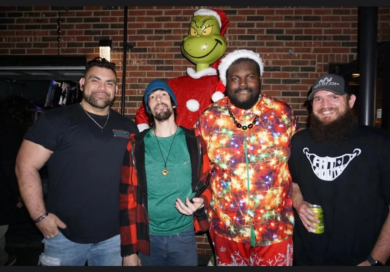 Four men posing in front of a brick wall, with a large Grinch figure wearing a Christmas Santa hat and sweater in the background. The men are wearing casual clothing, with some in Christmas-themed attire, smiling at the camera.