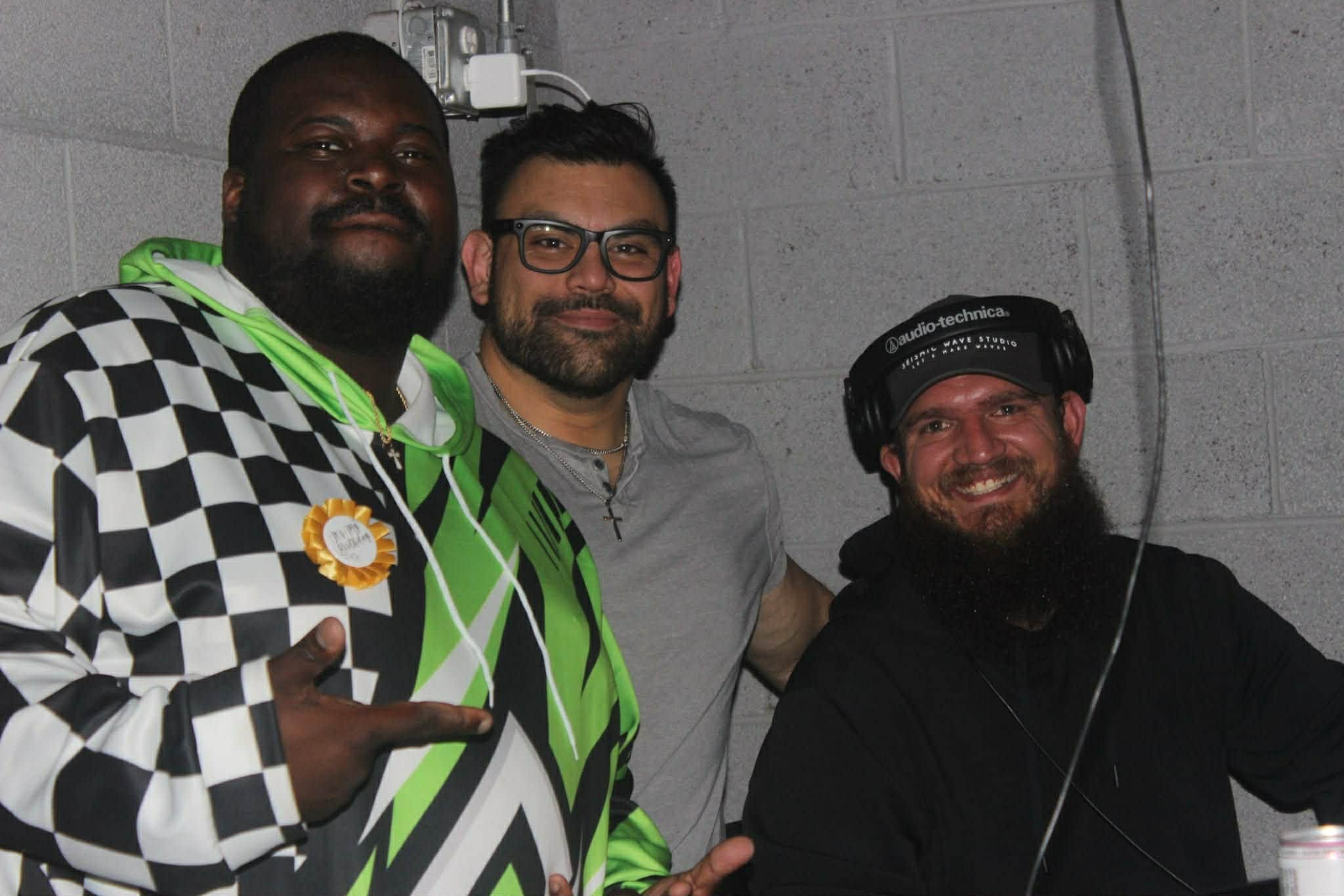 Three men smiling, one wearing a checkered black and white jacket with a ribbon badge, another in a gray shirt, and the third in a black hoodie with headphones, posing together against a gray cinder block wall.