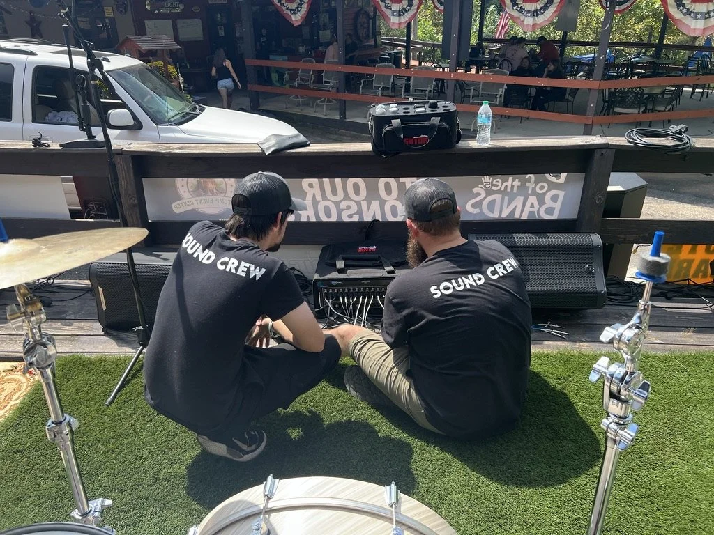 Two sound crew members sitting on the grass in front of a portable sound system, likely setting up or adjusting audio equipment. They are both wearing black t-shirts and black caps, with the words "SOUND CREW" printed on the back of their shirts. The scene appears to be outdoors at a venue with a stage, with tables, chairs, and a few people in the background, and patriotic banners hanging overhead.