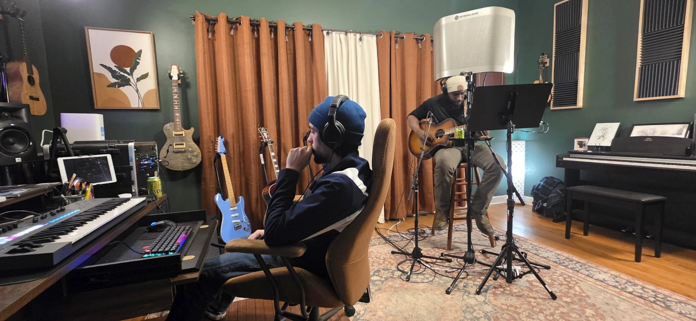 Two musicians in a recording studio, one sitting at a mixing desk with a computer and synthesizer, and the other playing an acoustic guitar in front of a microphone. The studio has green walls, wood flooring, and various guitars mounted on the wall.