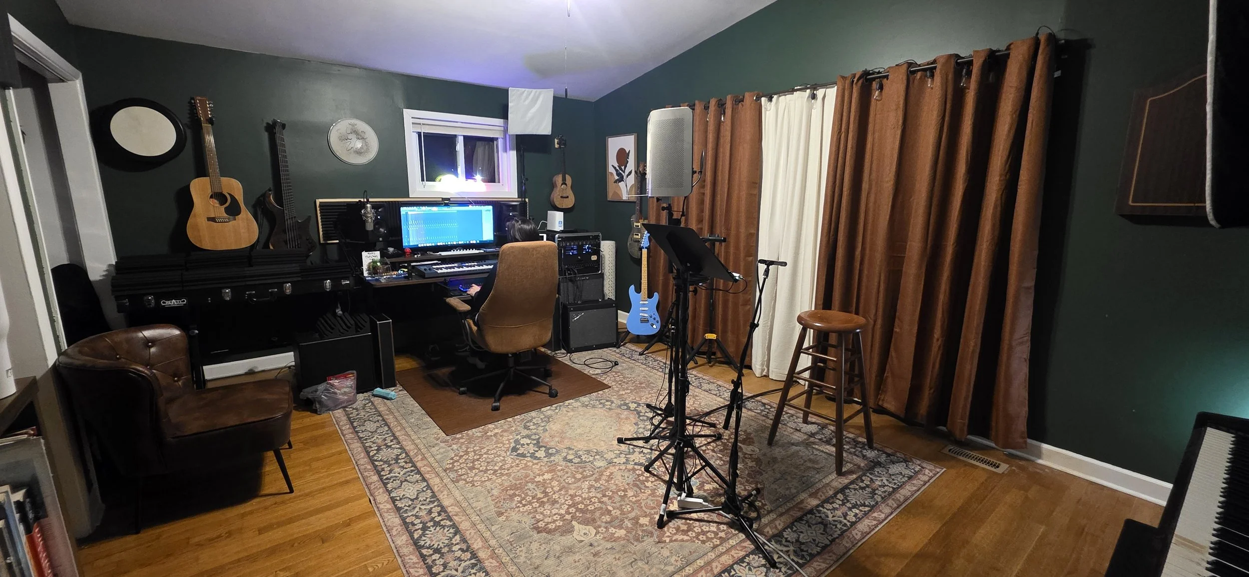 A music studio with guitars on the wall, a computer monitor, keyboard, microphone stands, and a rug. Curtains cover a window, and various musical equipment is present.