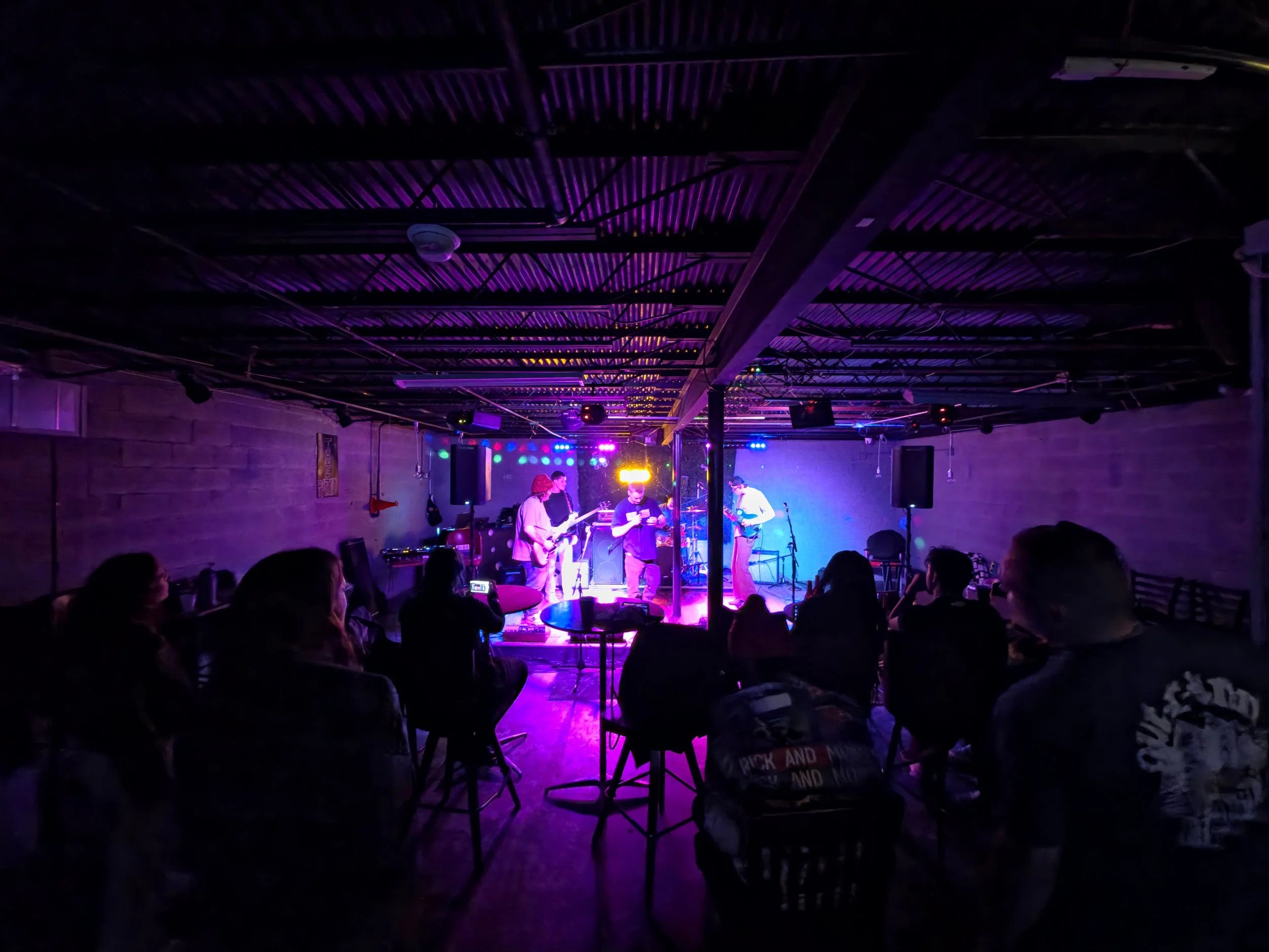 A live band performs on stage in a dimly lit venue with colorful lights, while an audience watches from tables and chairs.