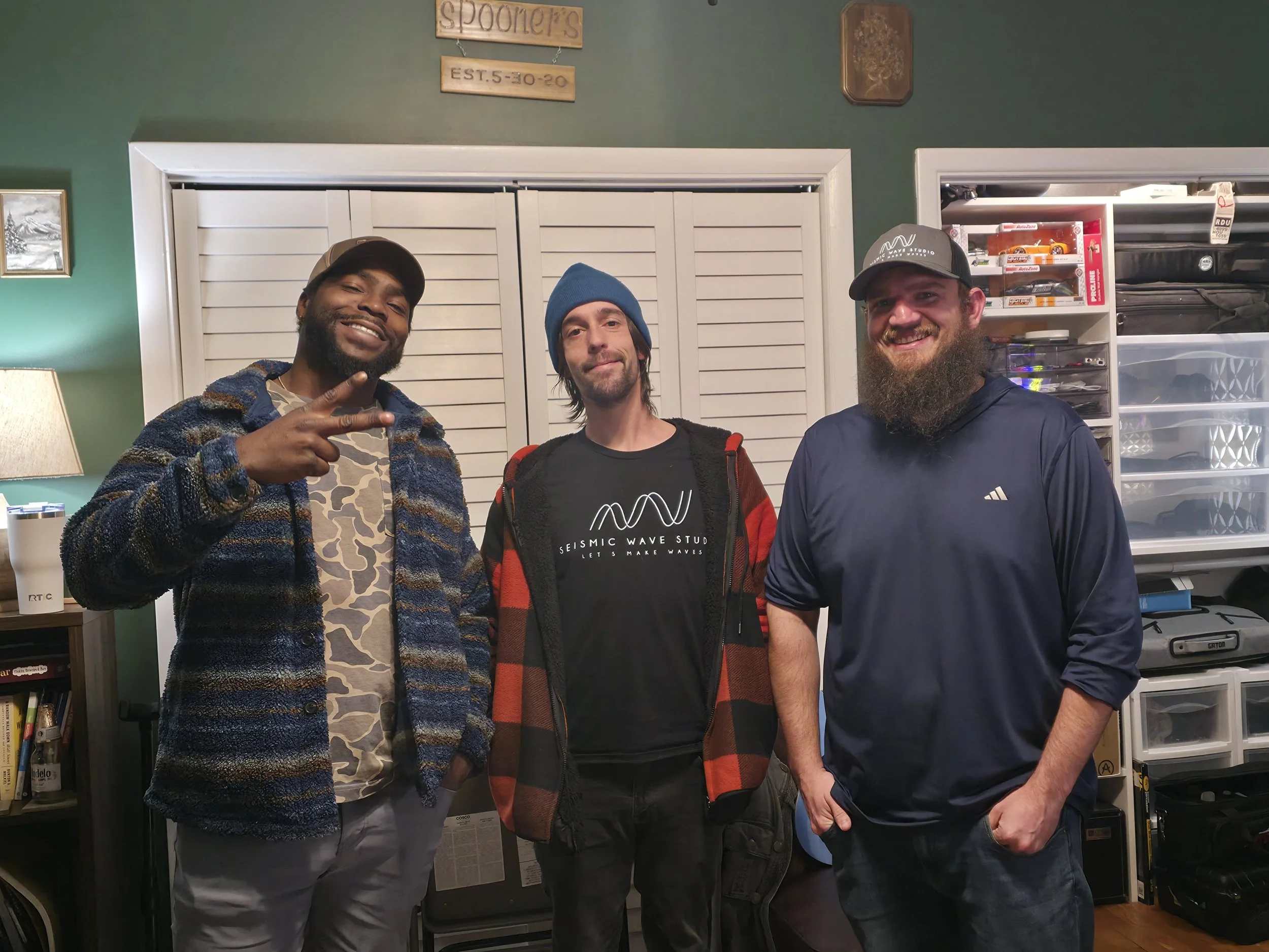Three men standing together inside a room, smiling and posing for the photo. The man on the left has dark skin, a beard, and is making a peace sign with his hand. The middle man has light skin, long hair, and is wearing a blue beanie and a black t-shirt with text. The man on the right has light skin, a beard, and is wearing a dark long sleeve shirt and a cap.