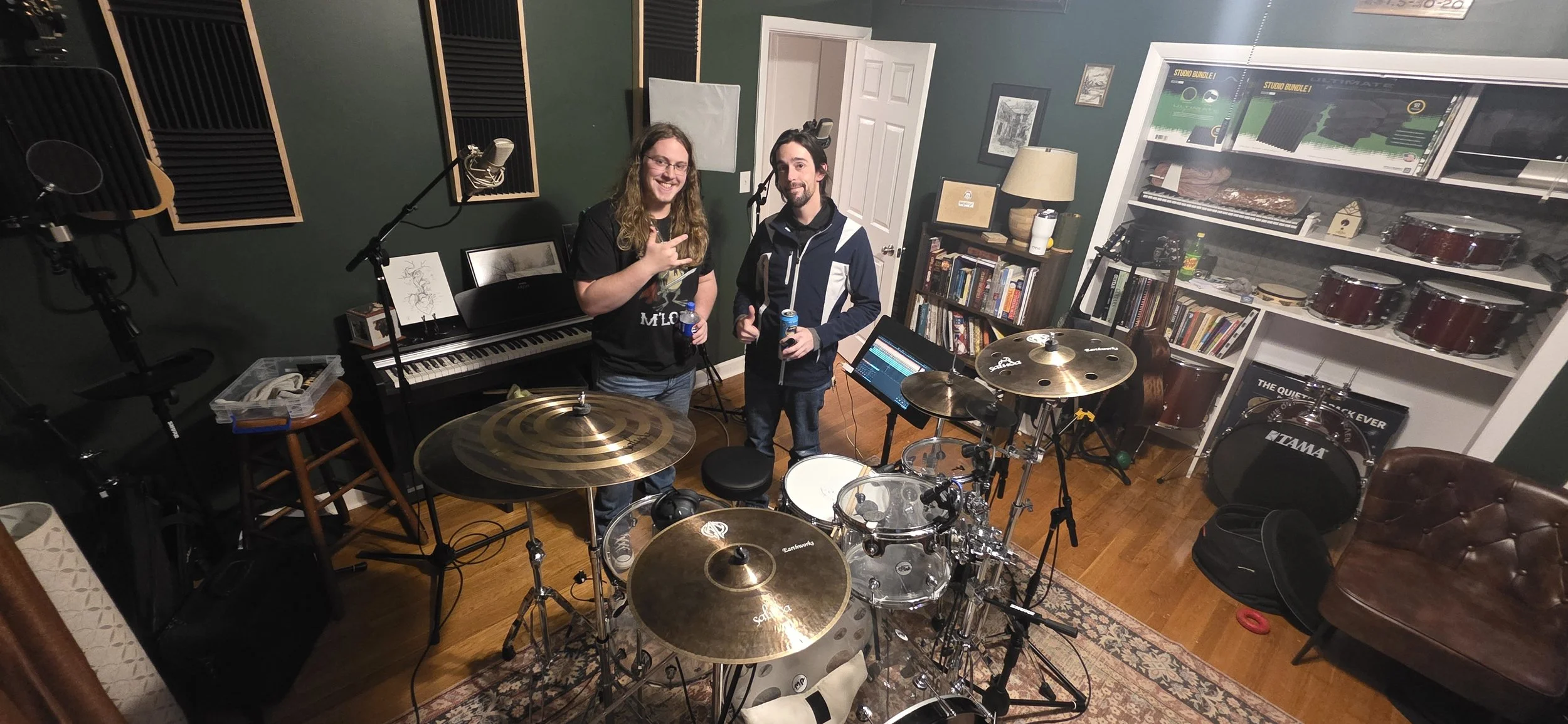 Two men in a music studio holding drinks, smiling, surrounded by musical instruments like drums, a keyboard, and microphones.