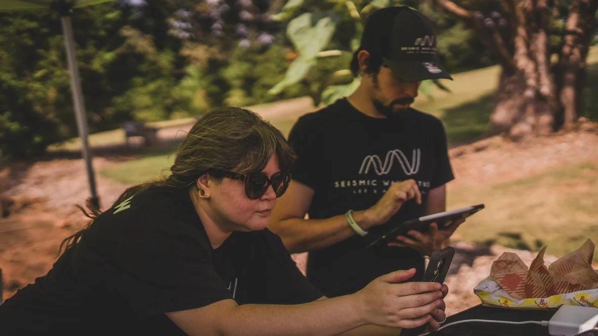 Two people sitting outdoors, one woman with sunglasses and long hair looking at her phone, and one man with a cap and beard using a tablet, with trees and a grassy area in the background.