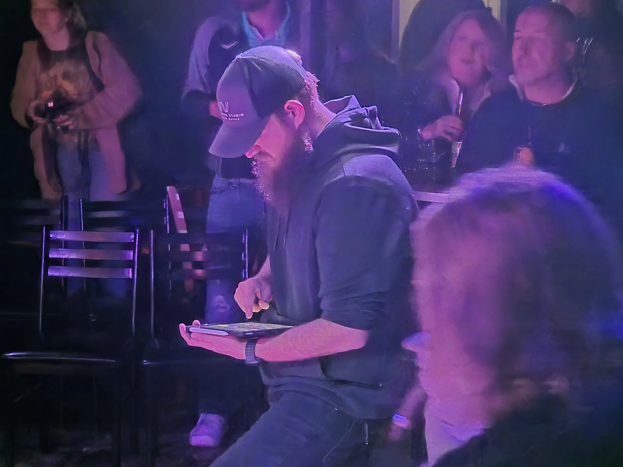 A man with a beard, wearing a cap and hoodie, is looking at a smartphone in a dimly lit venue, surrounded by several people, some of whom are drinking and some are taking photos.