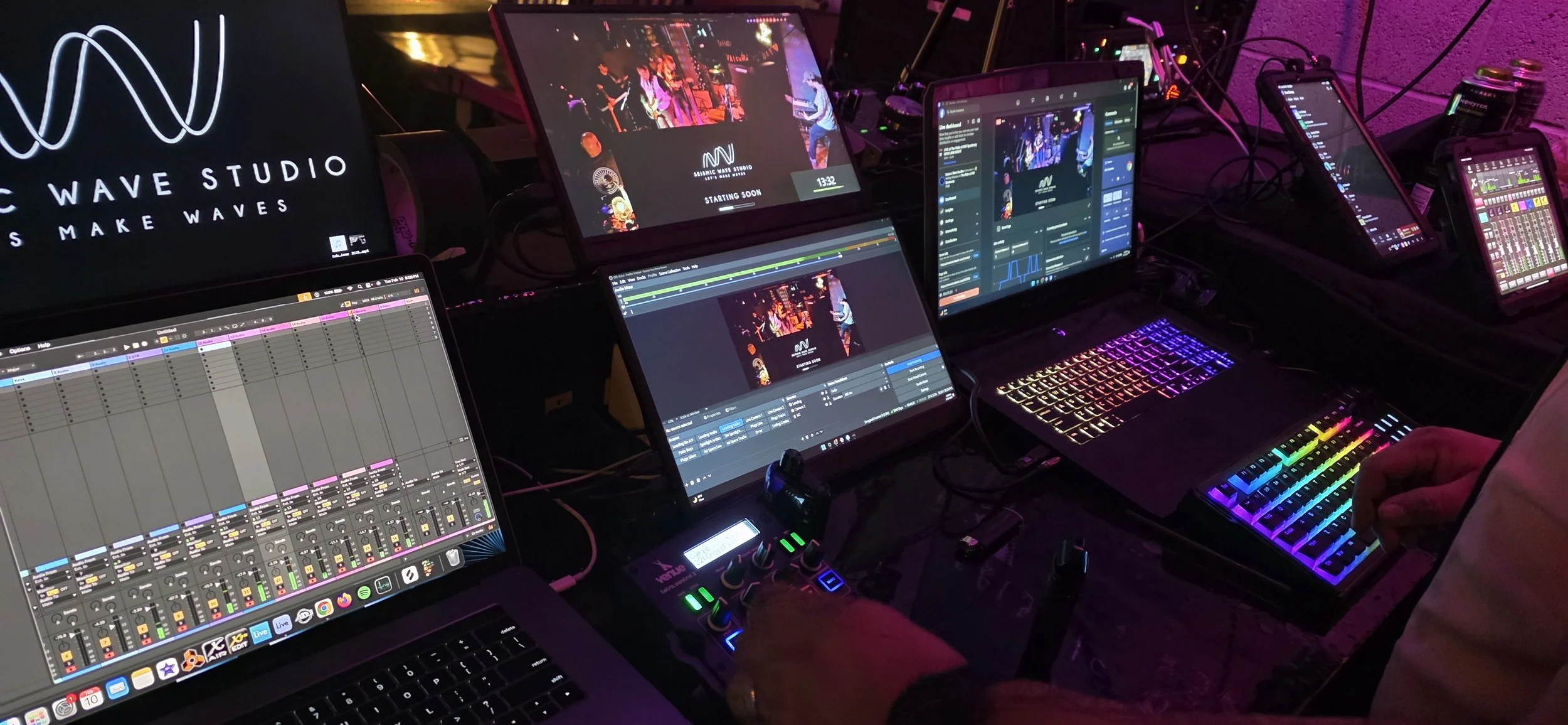 Multiple screens displaying music production software, waveforms, and a live music stream at Wave Studio with colorful backlit keyboards and DJ equipment in a dark room.