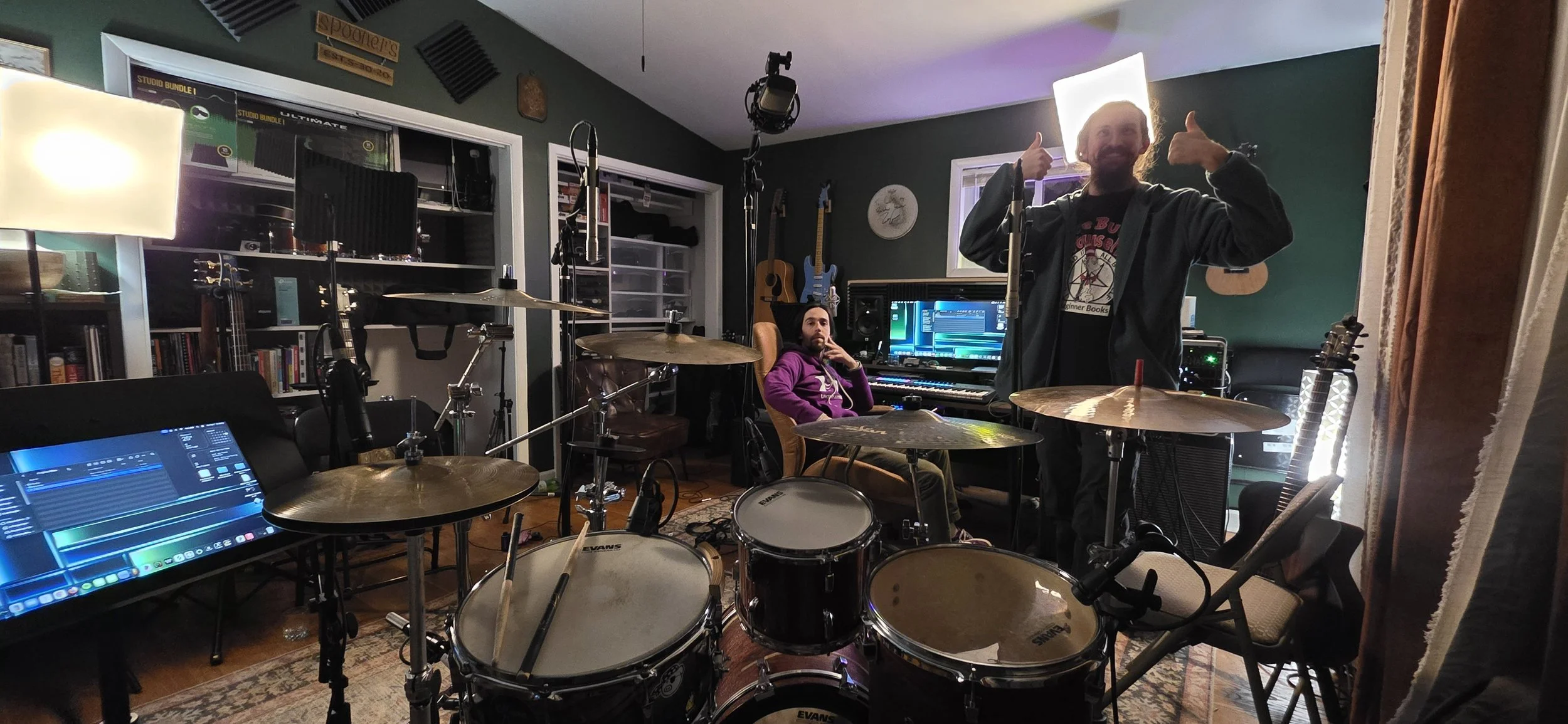A music studio with two men, one sitting at a digital piano and the other standing behind a drum set, giving a thumbs up. The room has guitars on the wall, a computer, and recording equipment.