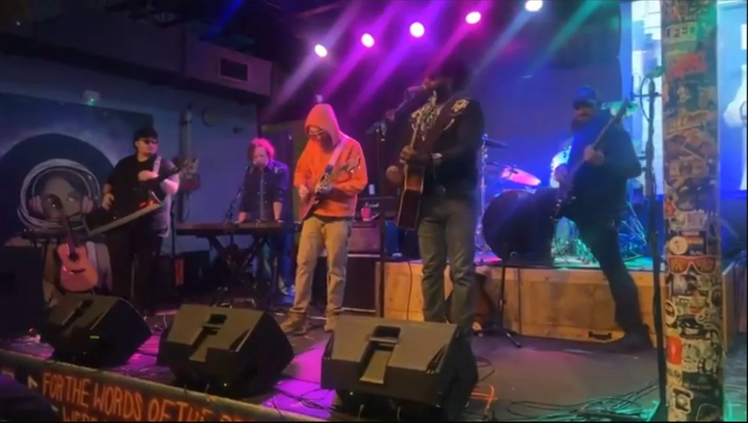 A live musical performance with five musicians on stage, including a keyboard player, a person in an orange hoodie playing guitar, a person in a black jacket with embroidery also playing guitar, a bassist, and a drummer in the background. The stage is lit with colorful purple and blue lights.