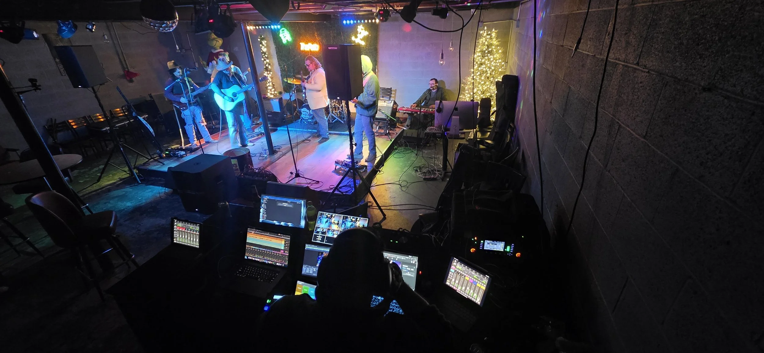 A live band performing on stage with Christmas decorations, including Christmas trees and Christmas lights, in a dimly lit venue with a sound engineer operating audio equipment.