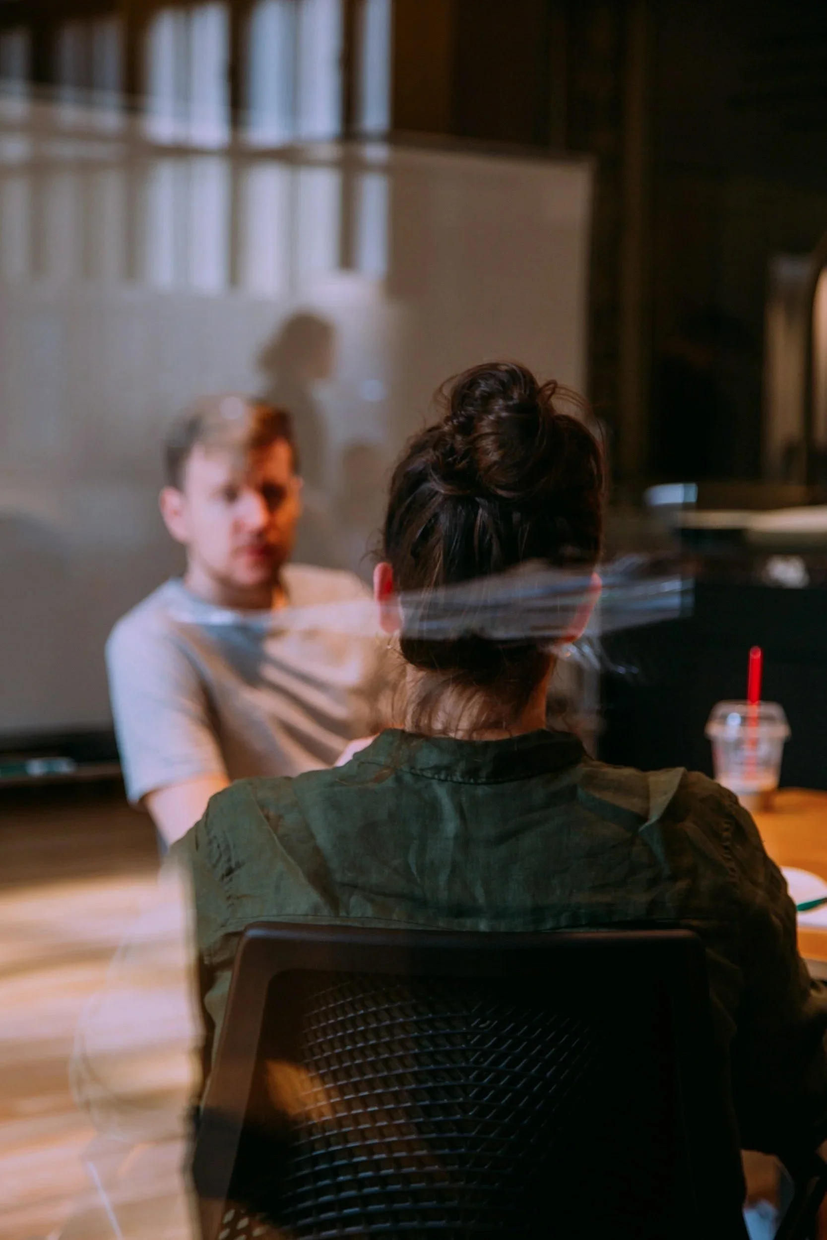 An athlete and a sport psychologist are sitting at a table engaged in a consultation, with a blurred man facing away and a woman with her hair in a bun, reflected on a glass surface, in a cozy indoor setting.