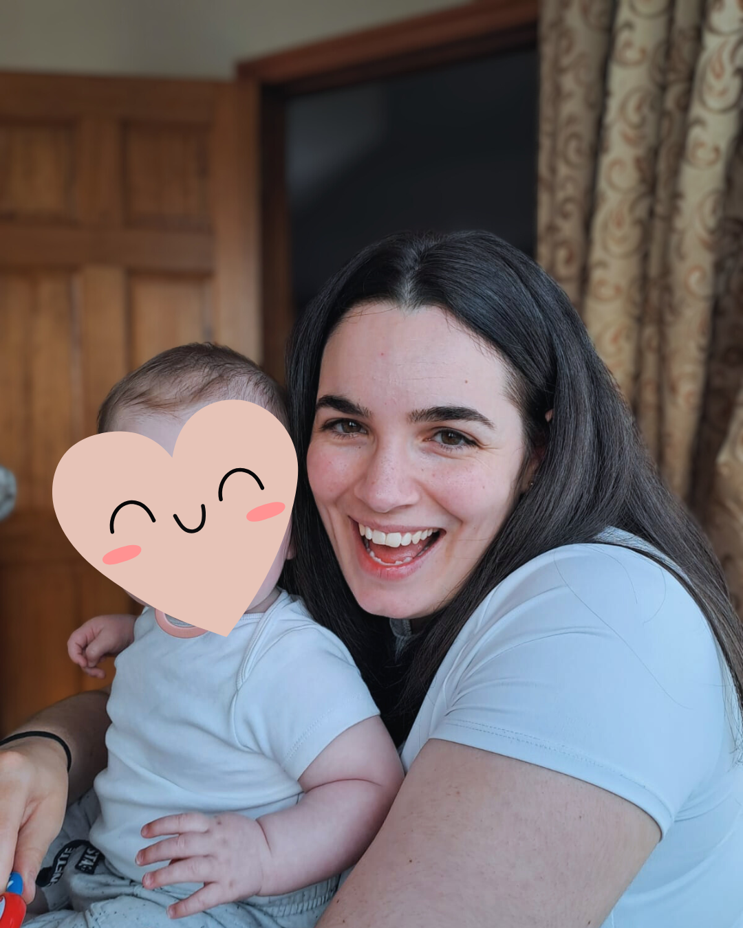 Smiling woman holding a baby with a heart face sticker in a room with wood-paneled walls and curtains.