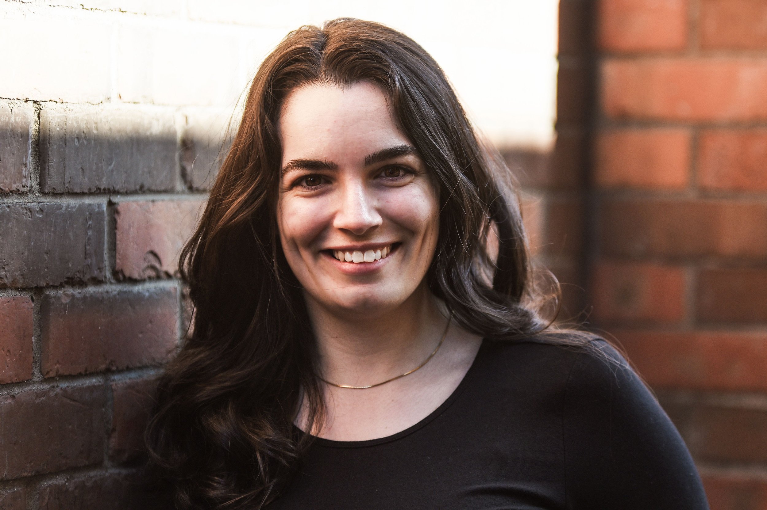 A smiling woman with dark brown hair, wearing a black top and a delicate necklace, standing against a brick wall outdoors.
