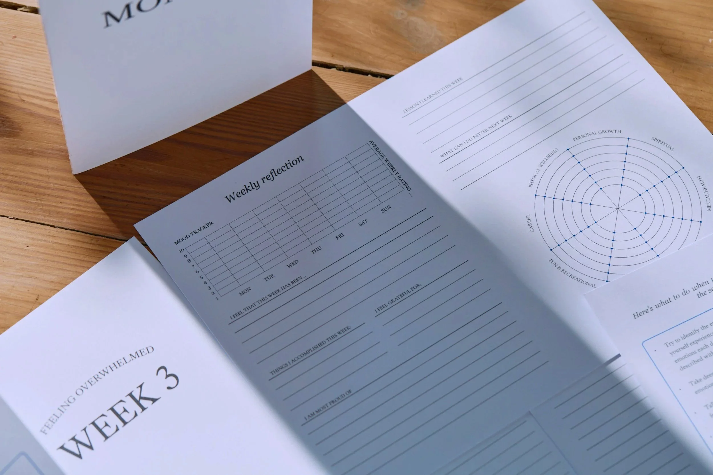 Open notebook with weekly reflection and mood tracker on a wooden table, sunlight casting shadows.