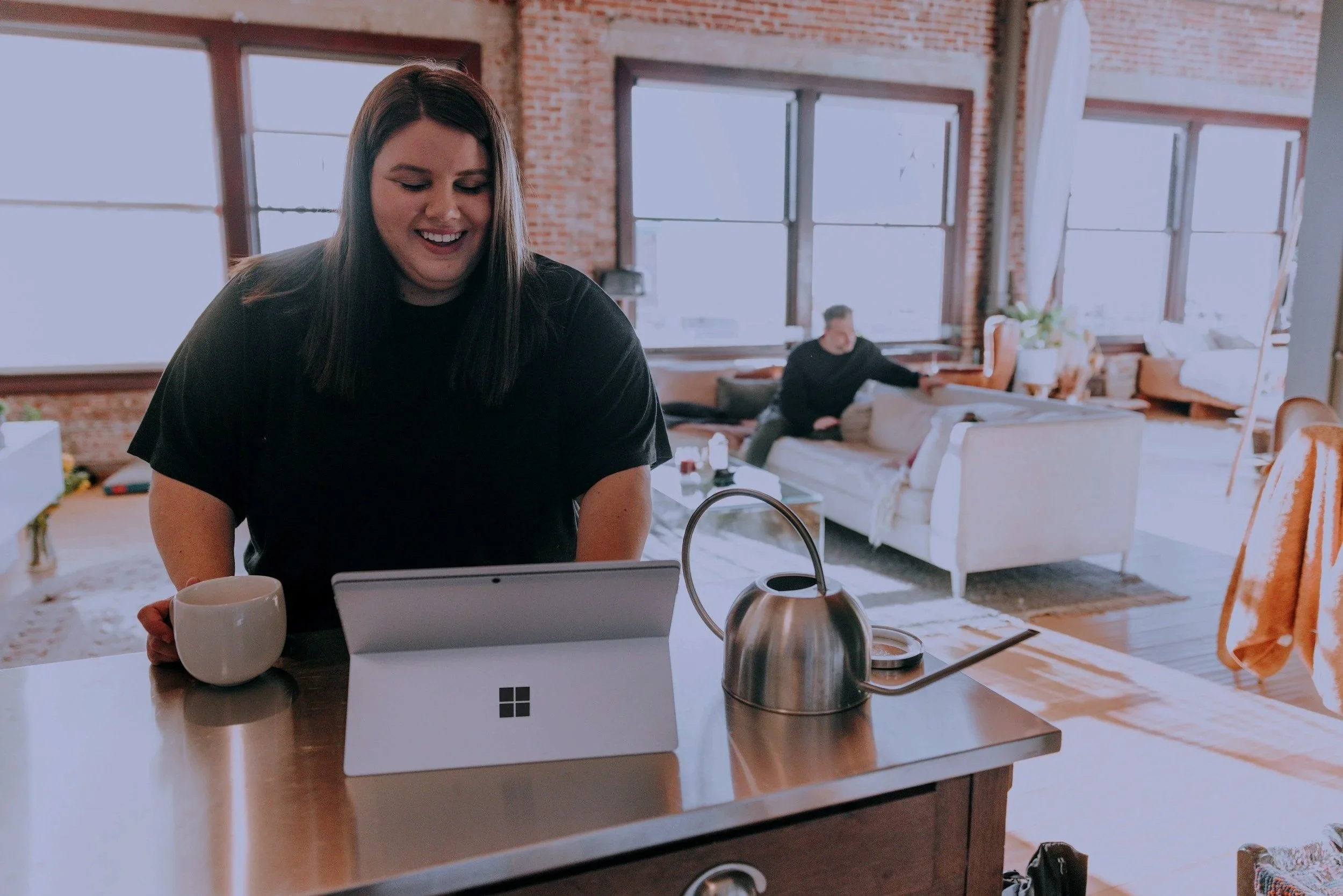 A woman smiling at a tablet on a kitchen counter with a coffee mug beside her, in a bright, modern living room with large windows.