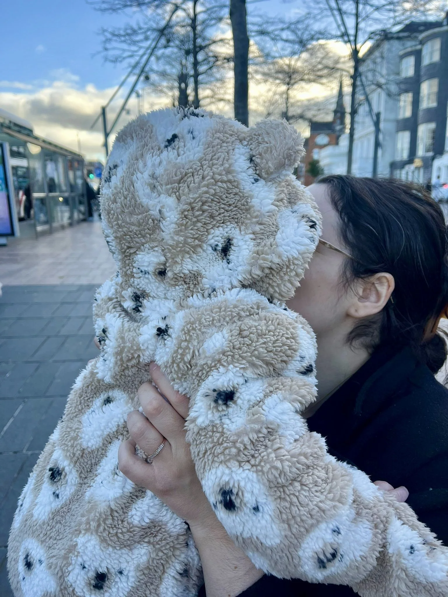 Person holding a baby in large fluffy teddy bear pram suit and kissing it on the face in an outdoor urban setting with trees, buildings, and a bus stop visible in the background.