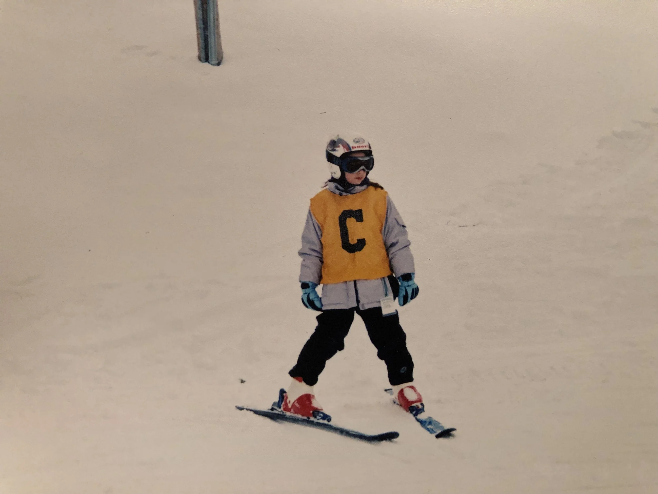 A young skier standing on snow, wearing a yellow vest with a black letter 'C', a helmet, goggles, gloves, jacket, and ski boots, with a ski pole in the background.