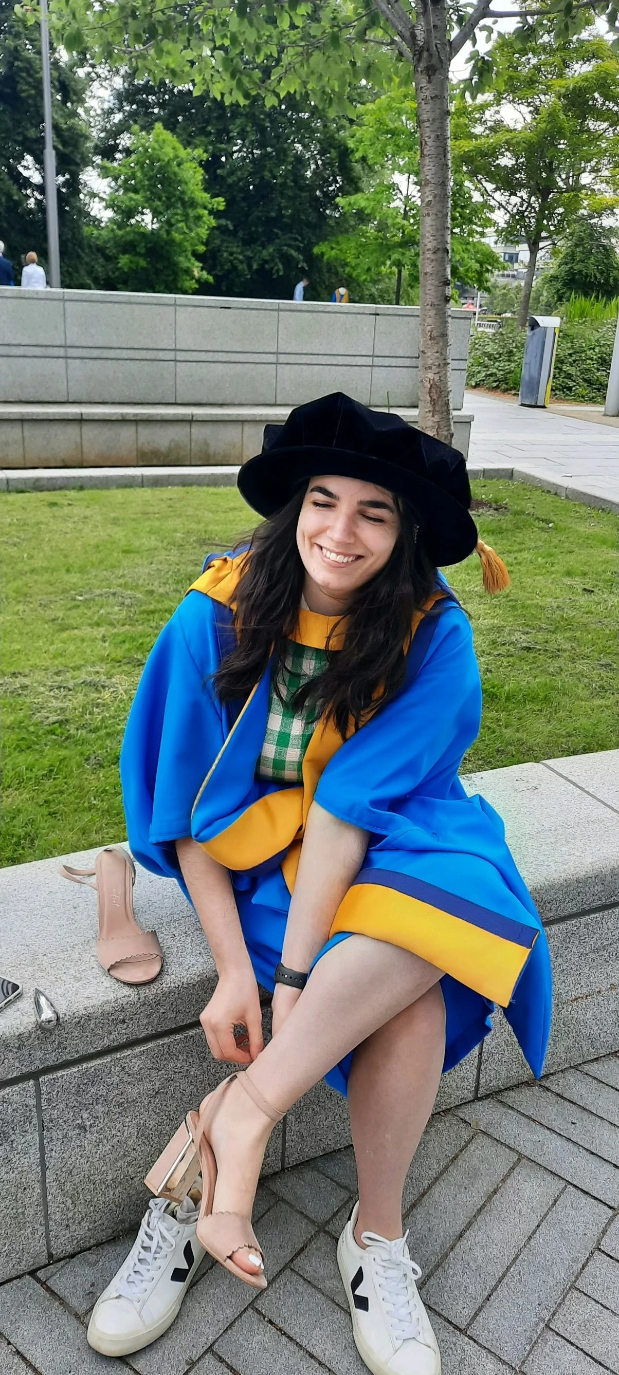 Sport psychologist Dr. Mckenzie Dow, a young woman in a blue and yellow graduation gown and cap, sitting on a stone bench outdoors, smiling with her eyes closed, with greenery and trees in the background.