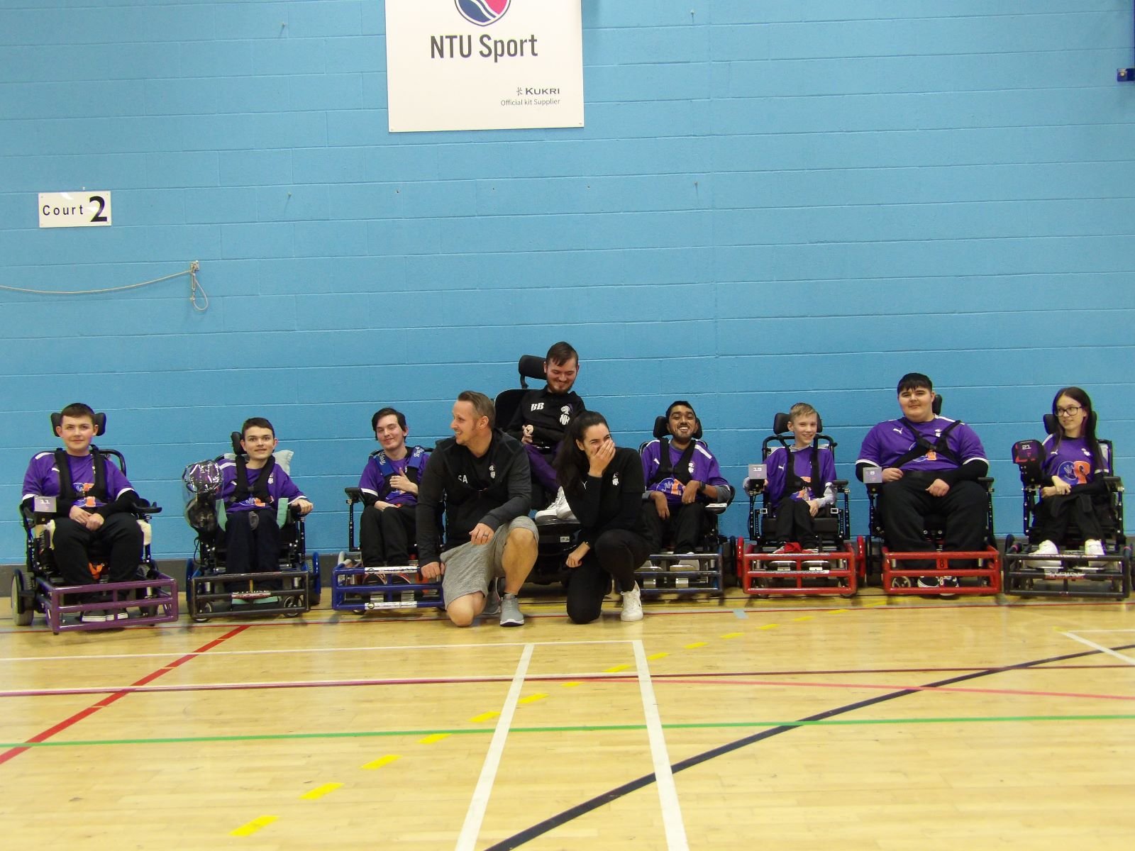 Sport psychologist Mckenzie Dow in a photo with a group of power wheelchair athletes and their coaches, posing for a photo in a gymnasium with blue walls, celebrating a sports event.