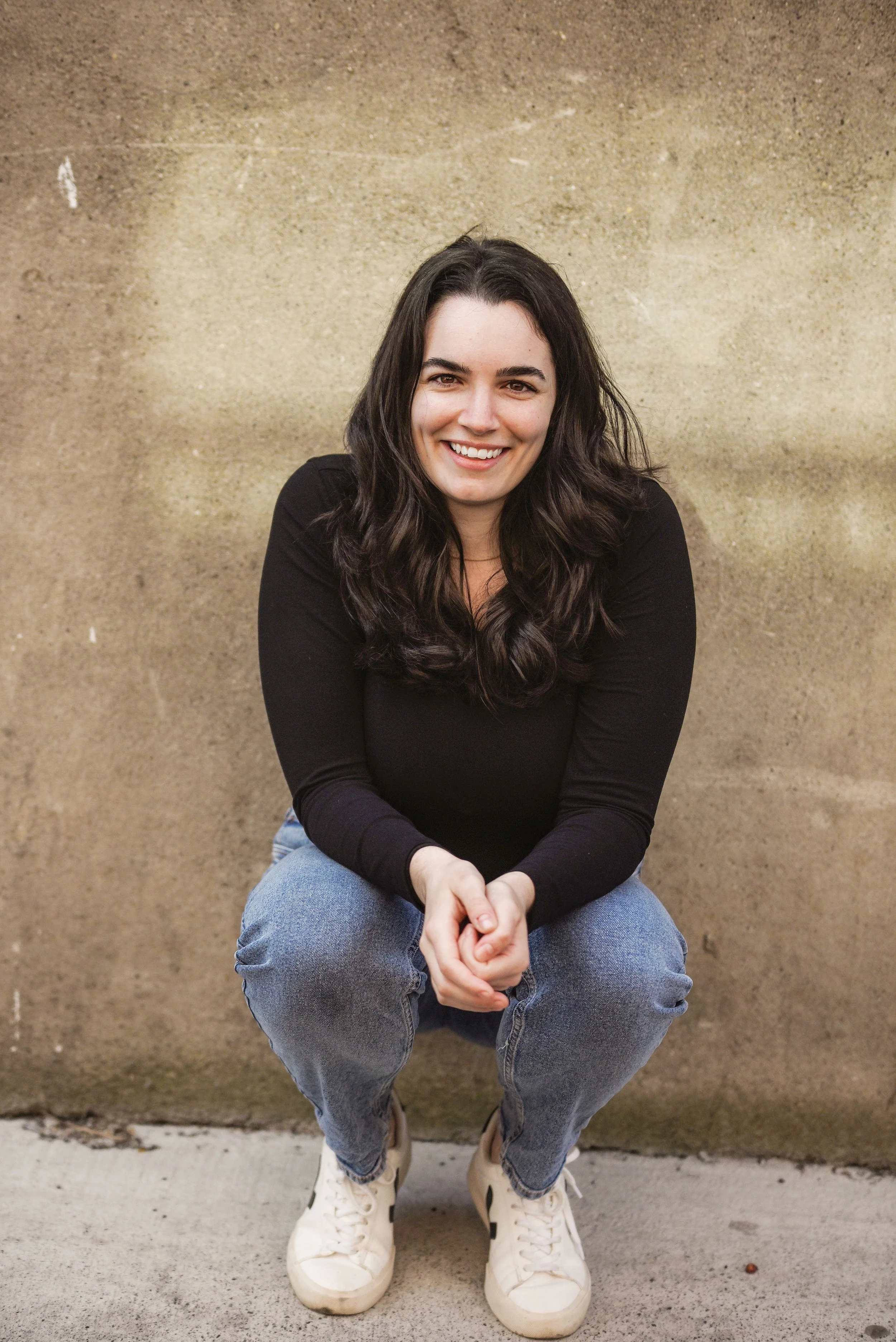 Sport Psychologist Mckenzie Dow, with dark wavy hair sitting in a squatting position against a beige wall, smiling at the camera, wearing a black top, jeans, and white sneakers.