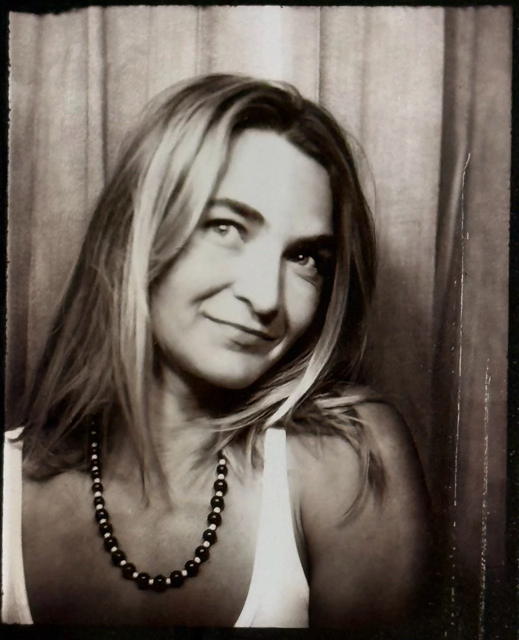 Black and white portrait of a woman with blonde hair wearing a white top and a beaded necklace, smiling slightly, against a dark curtain background.