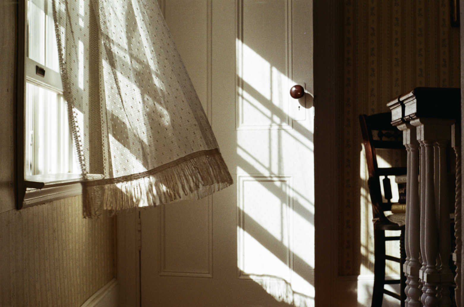 Interior of a room with sunlight streaming through a window with lace curtains, casting shadows on a white door and a chair with books.