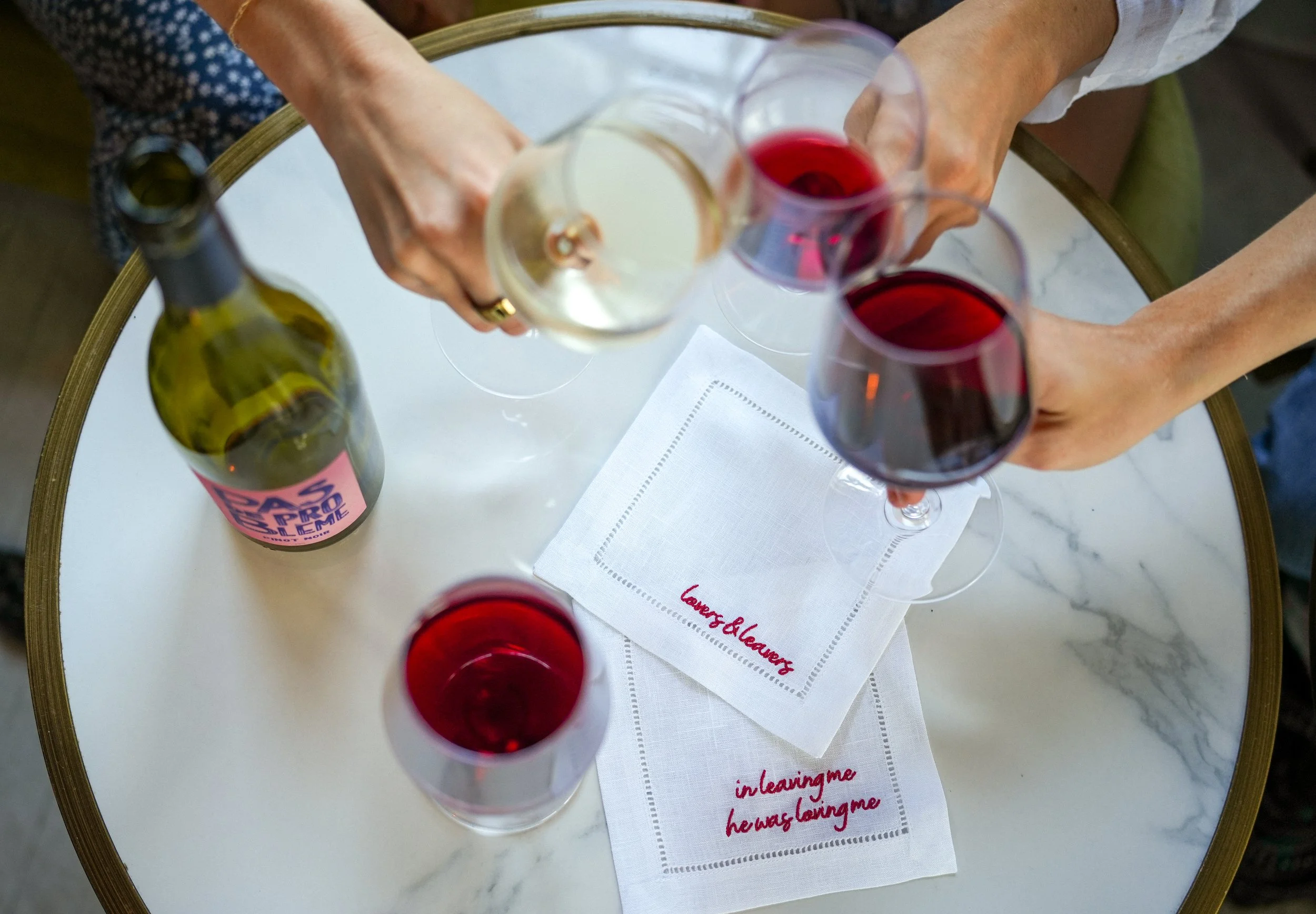 Overhead view of four glasses of wine on a round table, along with a wine bottle and a white cloth napkin with red script. Two glasses contain white wine, and two contain red wine.