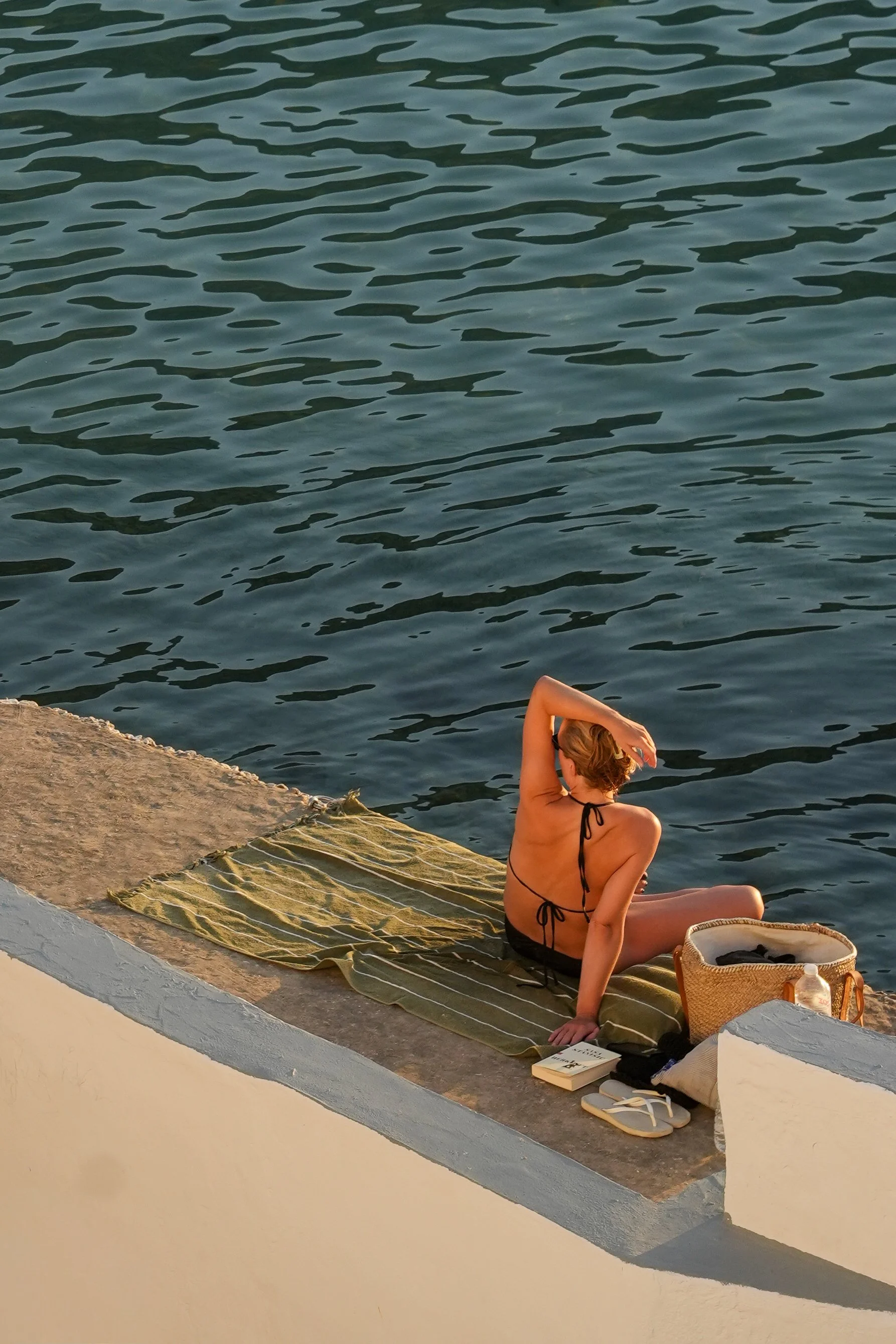 A woman in a black bikini sitting by the water on a striped towel, with a basket, books, and flip-flops nearby, overlooking a body of water with gentle ripples.