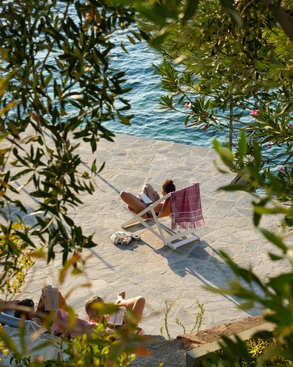 Person relaxing on a lounge chair by the water, reading a book, with a pink towel hanging on the chair, framed by green foliage.